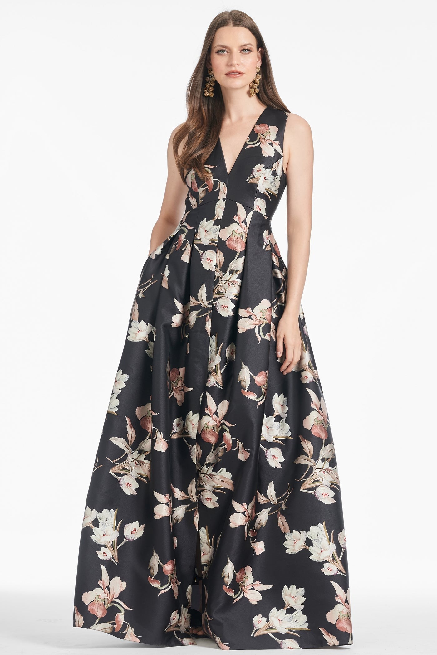 A woman stands confidently wearing the Sachin & Babi Brooke Gown - Tonal Petal. This elegant, floor-length black V-neck gown is adorned with a floral pattern featuring white and pink flowers, complete with box pleats and side pockets. She has long brown hair and wears dangling earrings, posing against a white background.