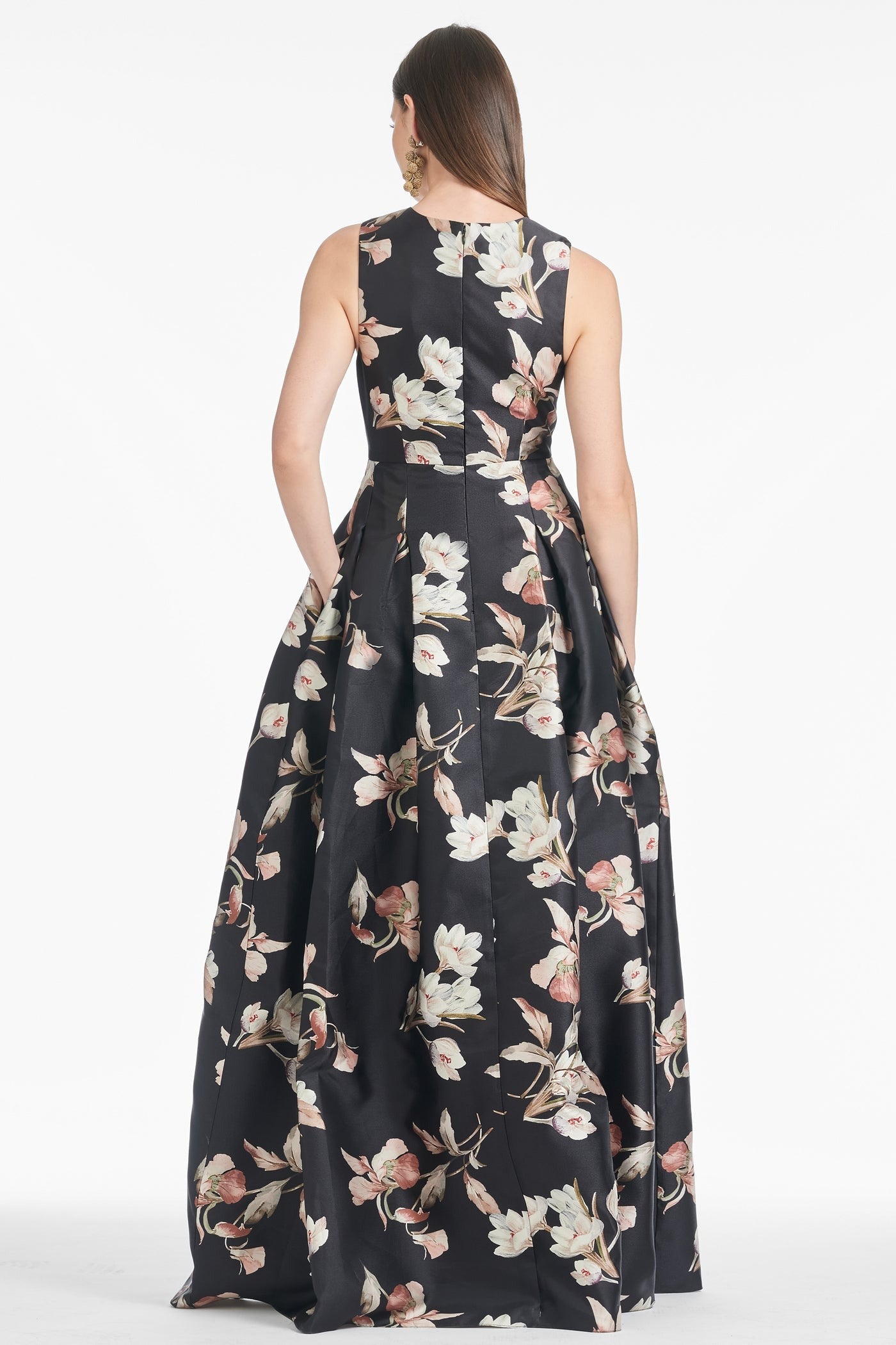 A woman is shown from the back wearing the Sachin & Babi Brooke Gown - Tonal Petal, a sleeveless, floor-length V-neck gown featuring a black base adorned with a white and pink floral pattern. The dress has a fitted bodice that flares into a full skirt with box pleats. She has long, straight hair and is standing against a plain white background.