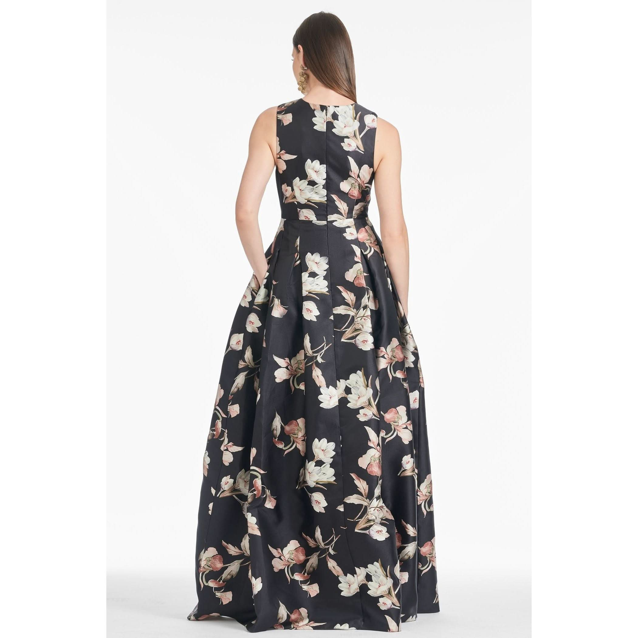 A woman is shown from the back wearing the Sachin & Babi Brooke Gown - Tonal Petal, a sleeveless, floor-length V-neck gown featuring a black base adorned with a white and pink floral pattern. The dress has a fitted bodice that flares into a full skirt with box pleats. She has long, straight hair and is standing against a plain white background.