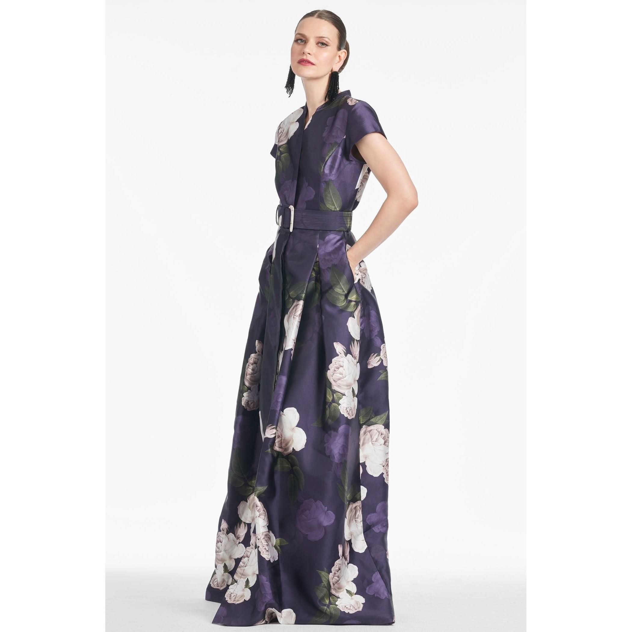 A woman is seen in the Sachin & Babi Marta Gown in Twilight Bloom, featuring a dark purple, floral pattern with box pleats and a belt. She wears earrings and has her hands in the gown's pockets while posing against a plain white background, gazing to the side.