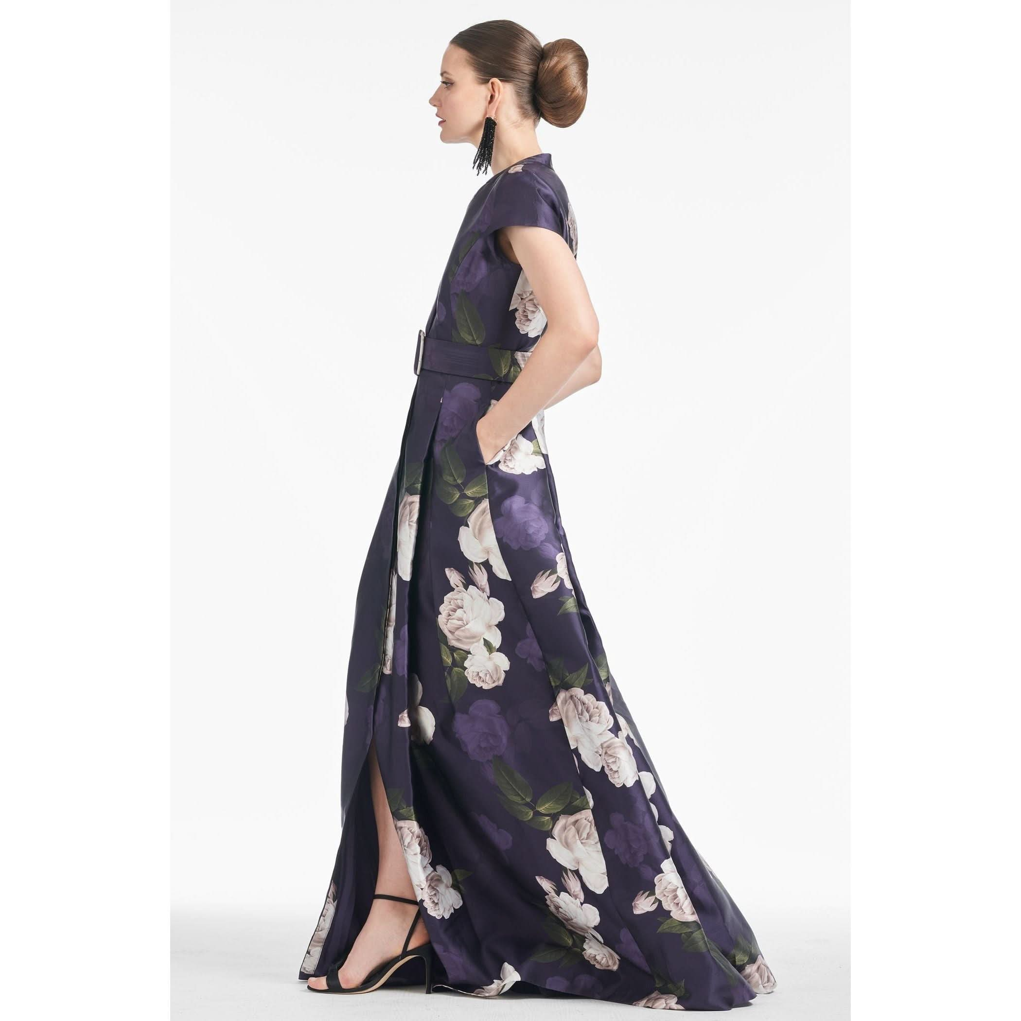 A woman in profile stands wearing the Sachin & Babi Marta Gown in Twilight Bloom, featuring a long, printed design with a belt. Her hair is styled in an elegant bun, and she wears dangly earrings alongside black high-heeled sandals. With one hand in her pocket, she exudes sophistication against the plain white backdrop.