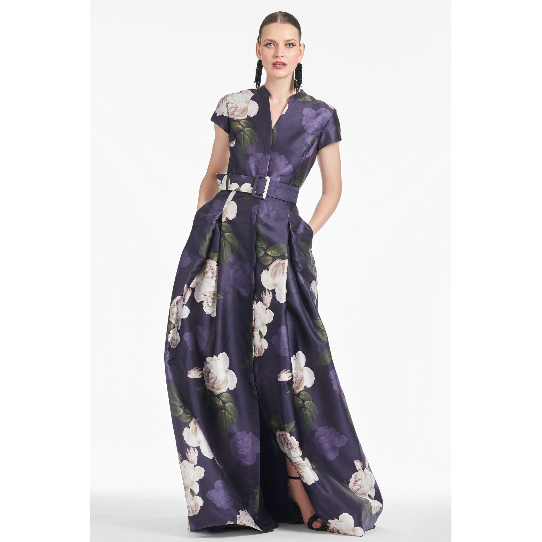 The individual exudes confidence in the Sachin & Babi Marta Gown in Twilight Bloom, showcasing a stunning dark purple V-neck adorned with large white floral patterns. This elegant dress features box pleats, a waist belt, and short sleeves. They accessorize with long earrings and sleekly pulled-back hair while looking directly at the camera.