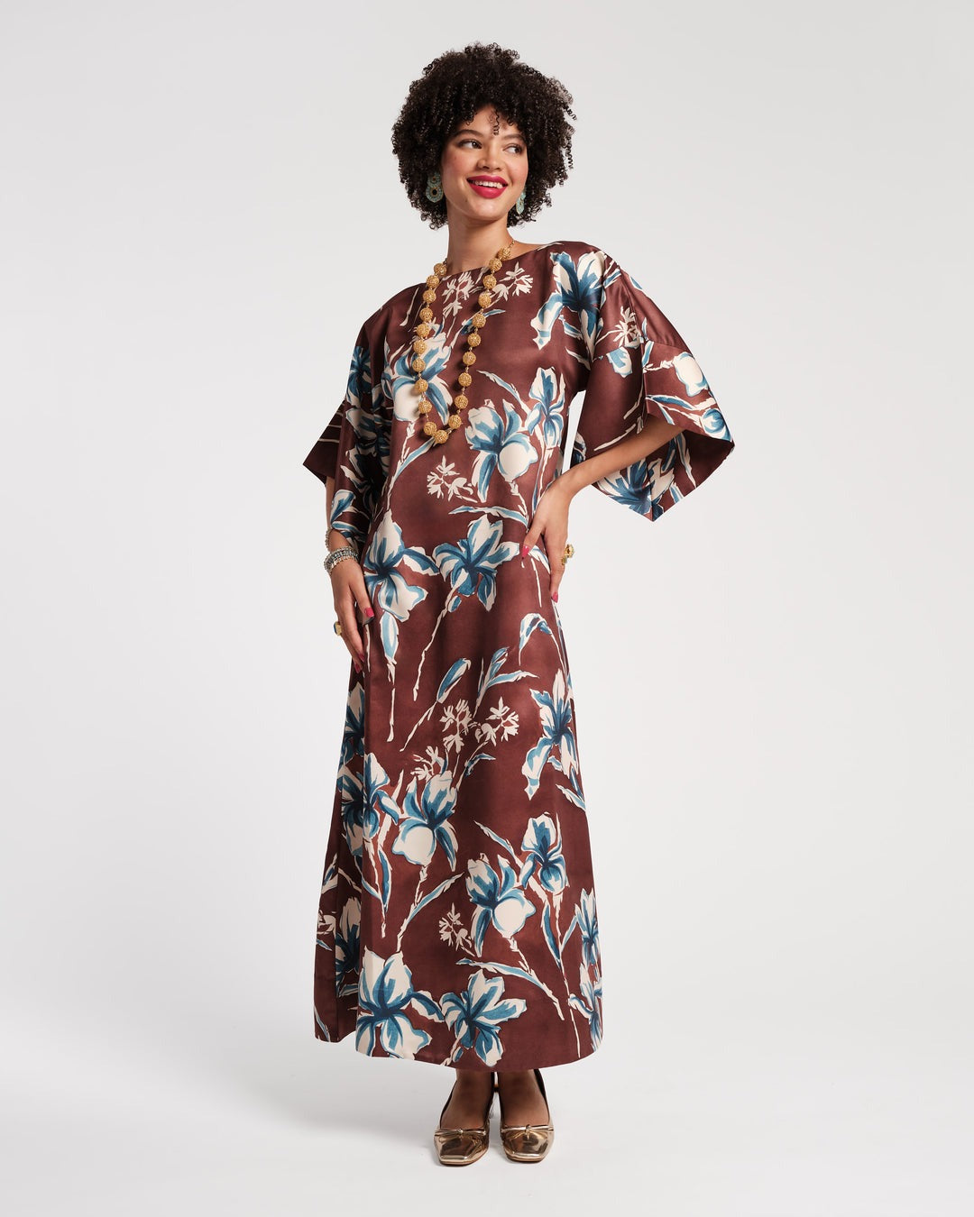 A woman with curly hair is wearing the Frances Valentine Spinnaker Maxi Dress in Lucky Devil Blue Iris, featuring a vintage-inspired tailored design with blue and white floral patterns on a brown background. She complements her outfit with a long gold bead necklace, matching earrings, and multiple rings. Posing confidently with one hand on her hip and the other resting by her side, she exudes timeless elegance.