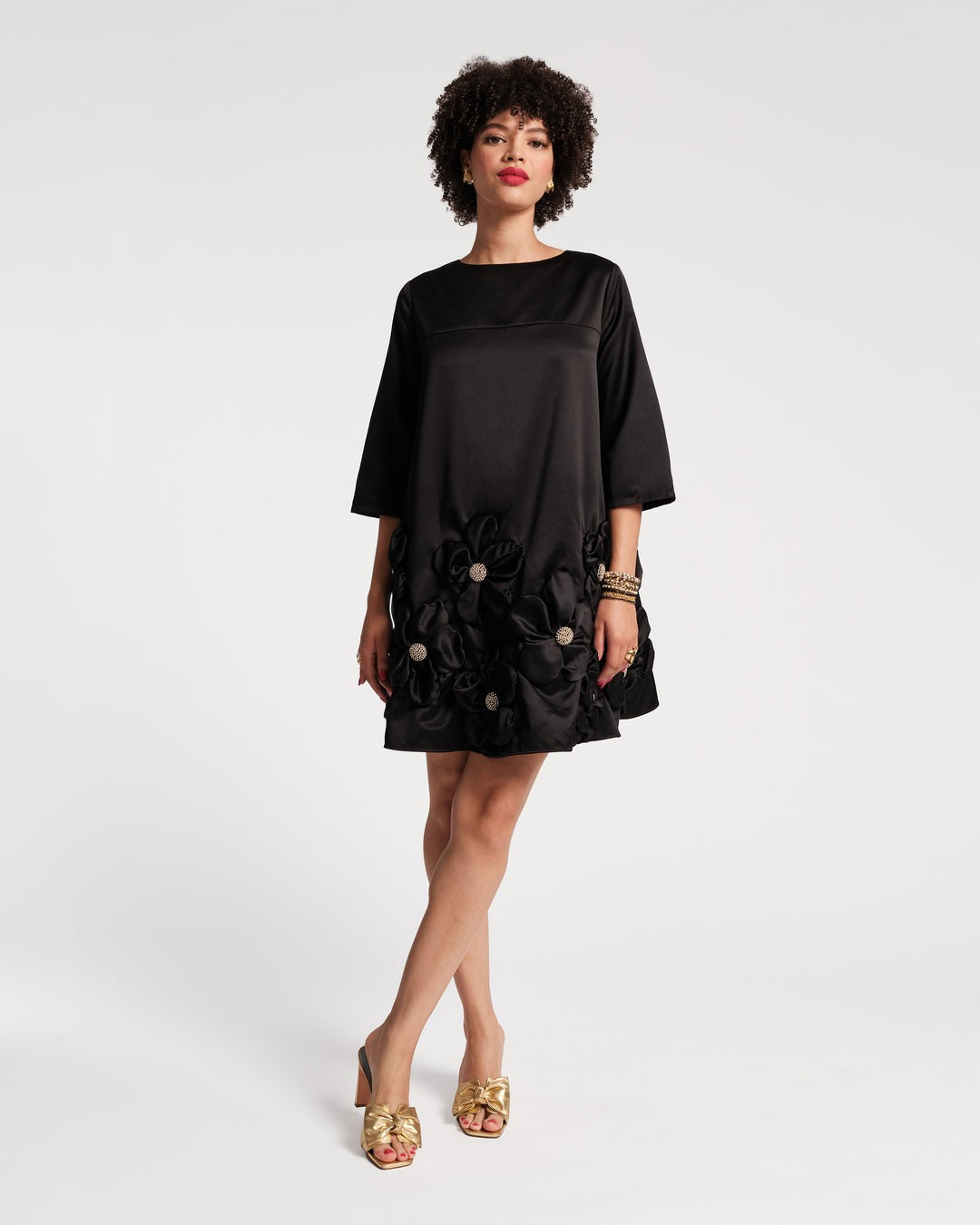 A woman in a Frances Valentine Edie Floral Mini Dress Black, adorned with 3D floral appliques at the hem, holds a small gold-textured clutch. She has curly hair and wears earrings with metallic beaded detail. Against a simple, light-colored background, she looks confidently at the camera.