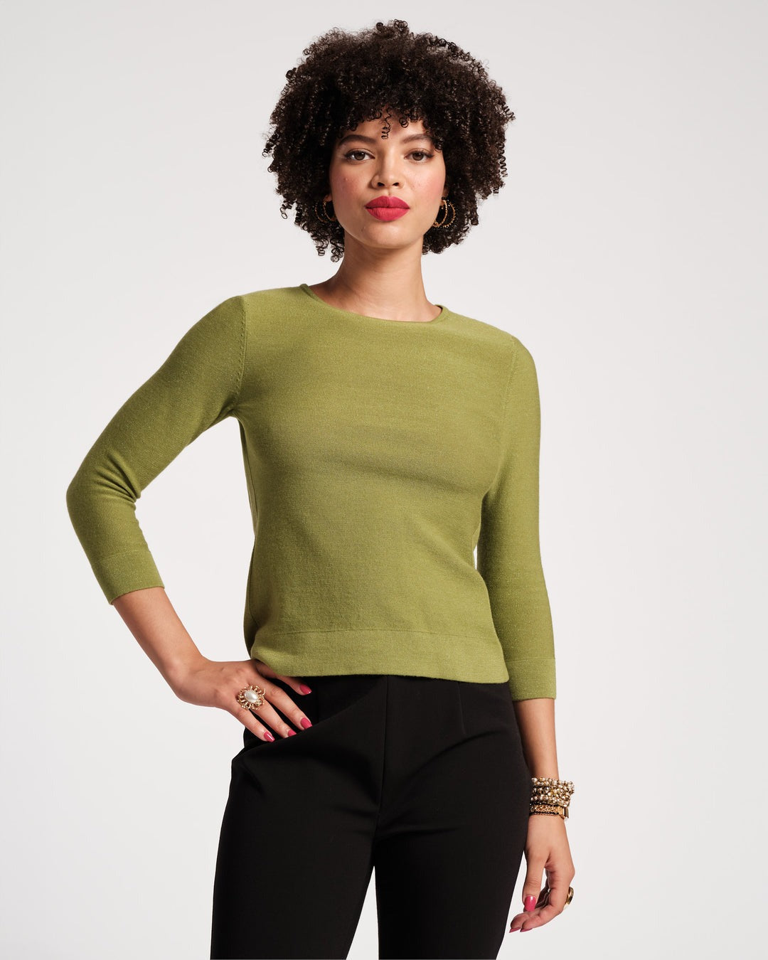 A person with short, curly hair is shown from the back, wearing a Frances Valentine Rachel Knit Top in Celery and black pants. The background is plain and light-colored. The person's left wrist is adorned with multiple bracelets, exemplifying a stylish fall wardrobe.