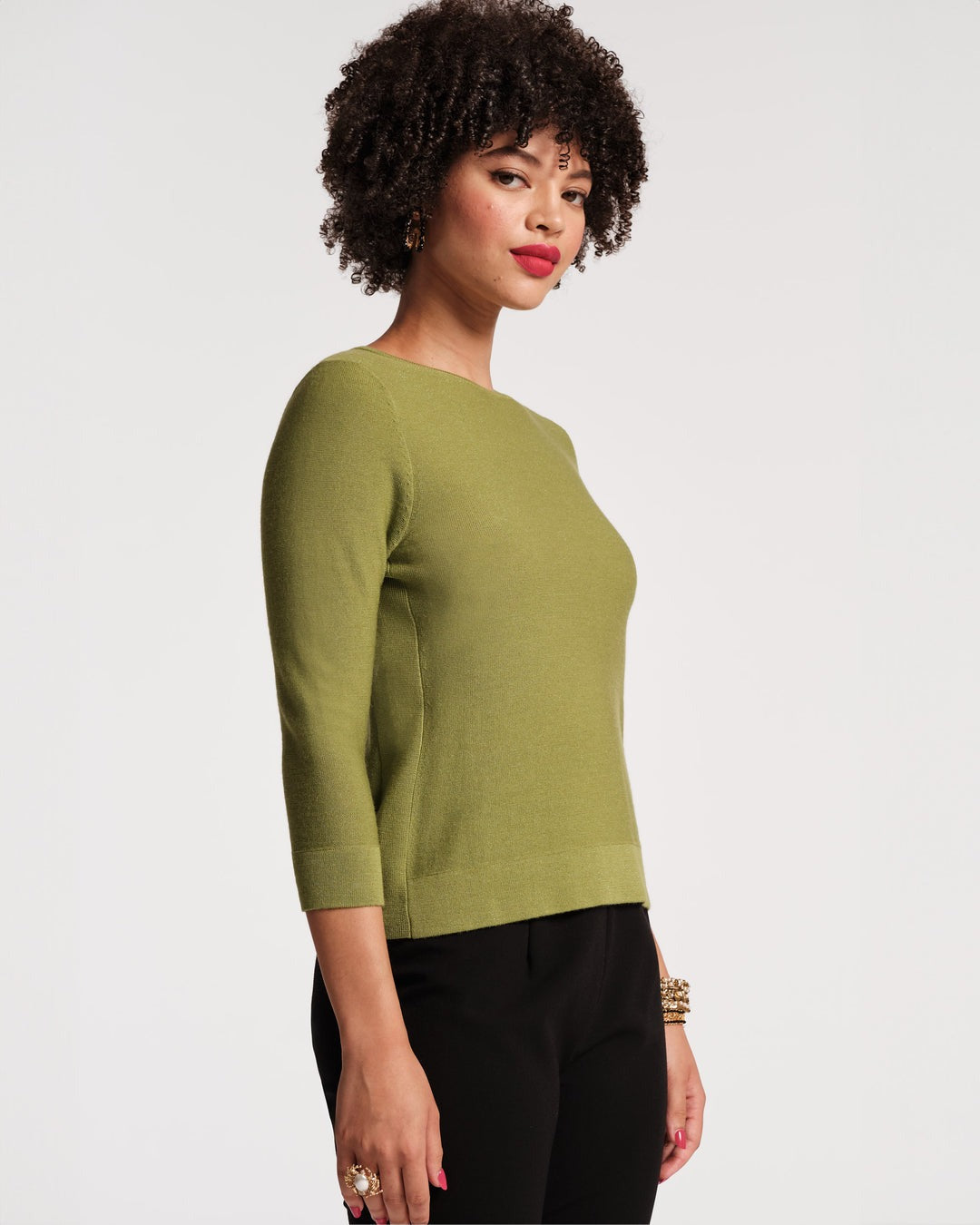 A person with short, curly hair is shown from the back, wearing a Frances Valentine Rachel Knit Top in Celery and black pants. The background is plain and light-colored. The person's left wrist is adorned with multiple bracelets, exemplifying a stylish fall wardrobe.