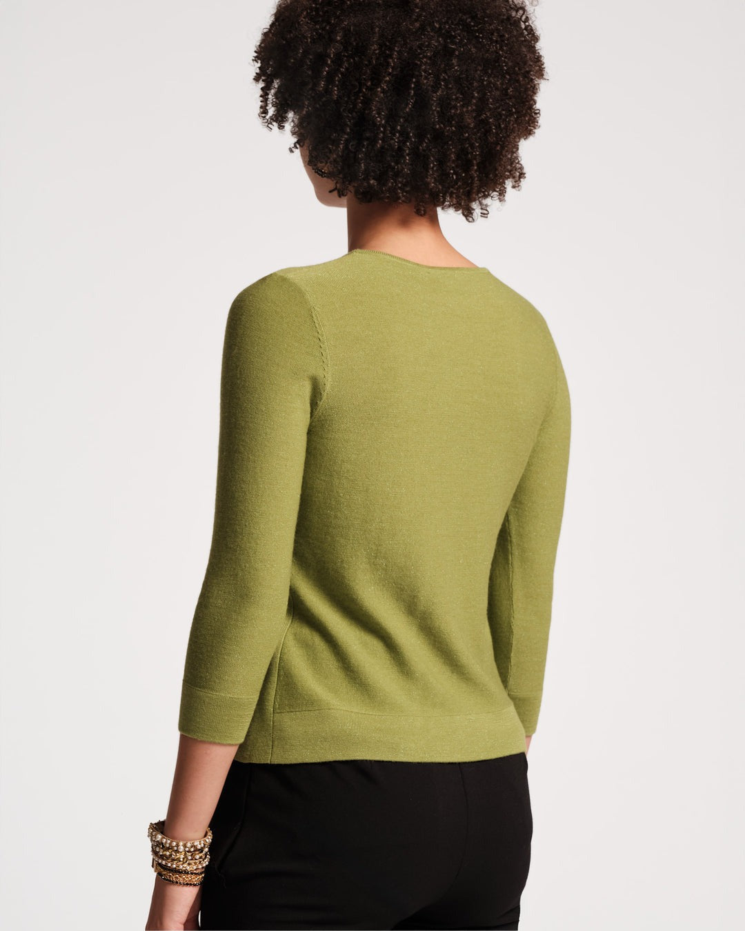 A person with short, curly hair is shown from the back, wearing a Frances Valentine Rachel Knit Top in Celery and black pants. The background is plain and light-colored. The person's left wrist is adorned with multiple bracelets, exemplifying a stylish fall wardrobe.