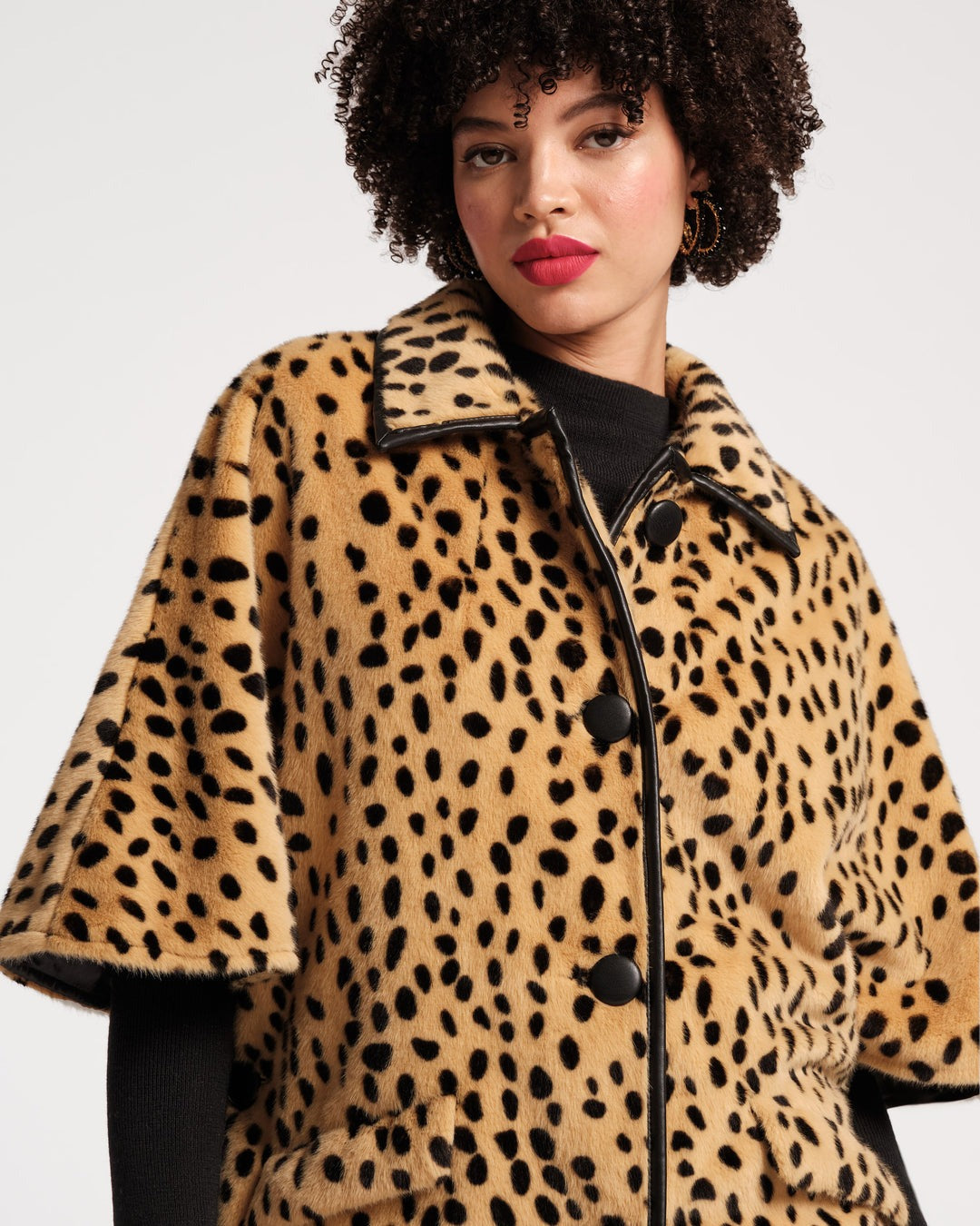 Close-up of a person wearing the stunning Frances Valentine Nico Poncho Faux Fur Cheetah Print coat, paired with a vintage-inspired ensemble that includes a black long-sleeved top. They showcase a gold ring with a large white stone on their left hand, accentuated by pink nail polish on their fingernails.