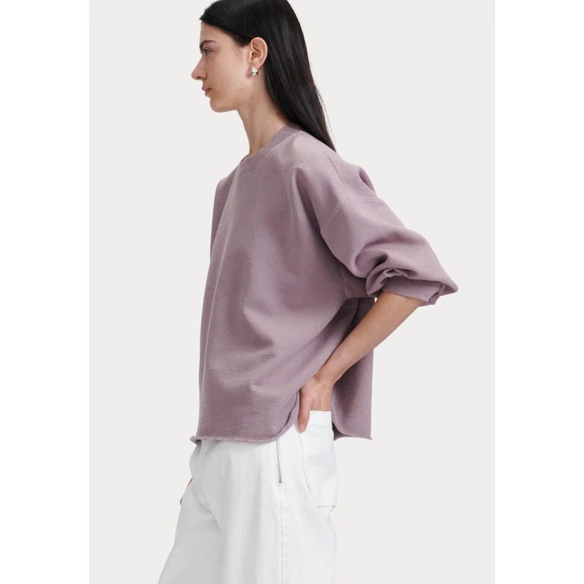 A person with long dark hair is shown in profile, wearing the Rachel Comey Fond Sweatshirt in mauve and white pants. Their right hand is casually placed in their pocket against a plain off-white background.