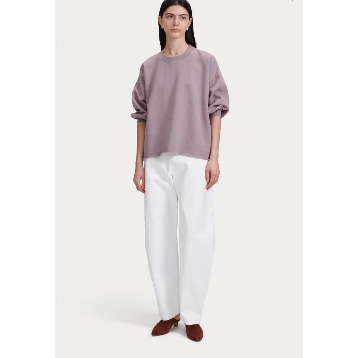 A person with long dark hair is wearing the Rachel Comey Fond Sweatshirt in mauve, paired with white wide-legged pants and brown shoes, standing against a plain background.