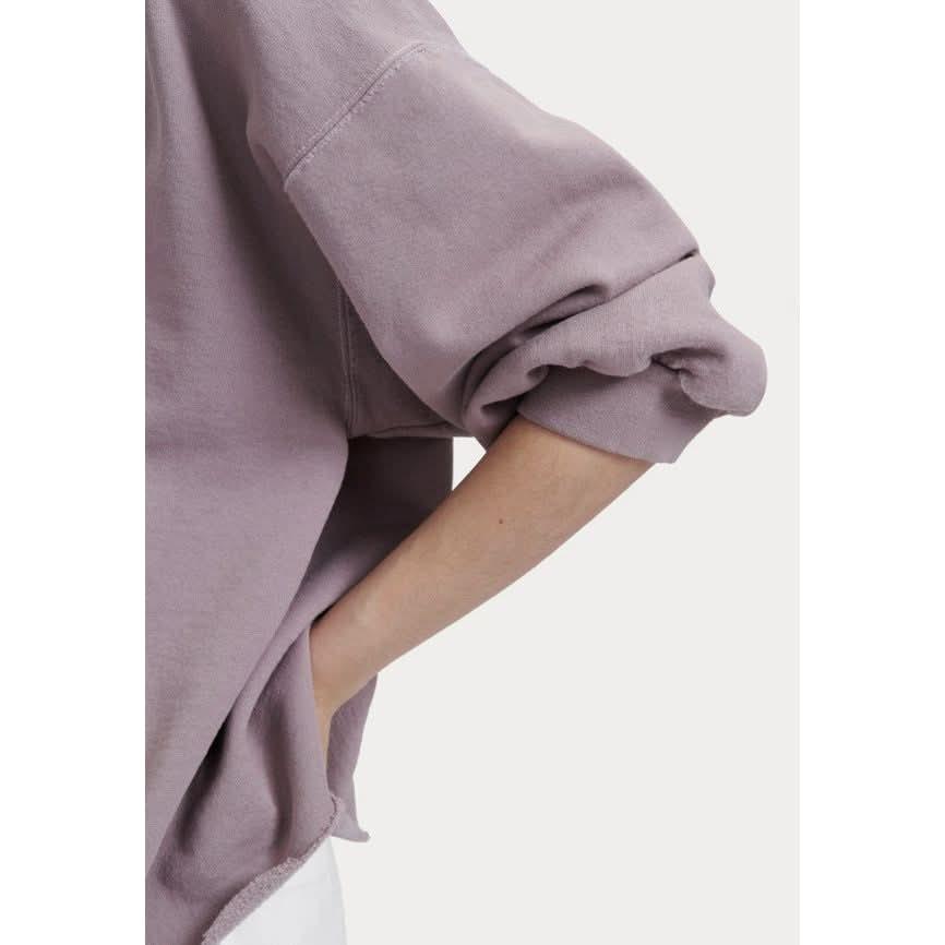 A person wearing the Rachel Comey Fond Sweatshirt in mauve, featuring its loose fit and balloon sleeves, with one hand casually tucked into a pocket of white pants. The attention is drawn to the ribbed knit pattern of the fabric, set against a plain background.