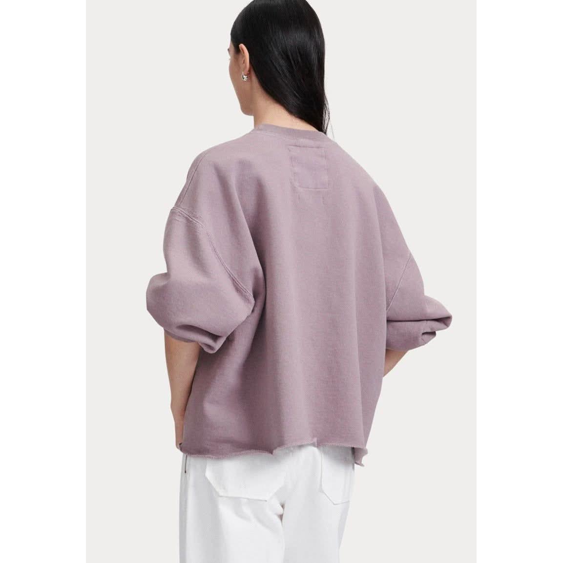 A person with long dark hair stands with their back to the camera, wearing a Rachel Comey Fond Sweatshirt in mauve, which is loose-fitting and features balloon sleeves. They pair it with white pants, and the sweatshirt's chunky knit cotton-like bottom has a slightly frayed edge.