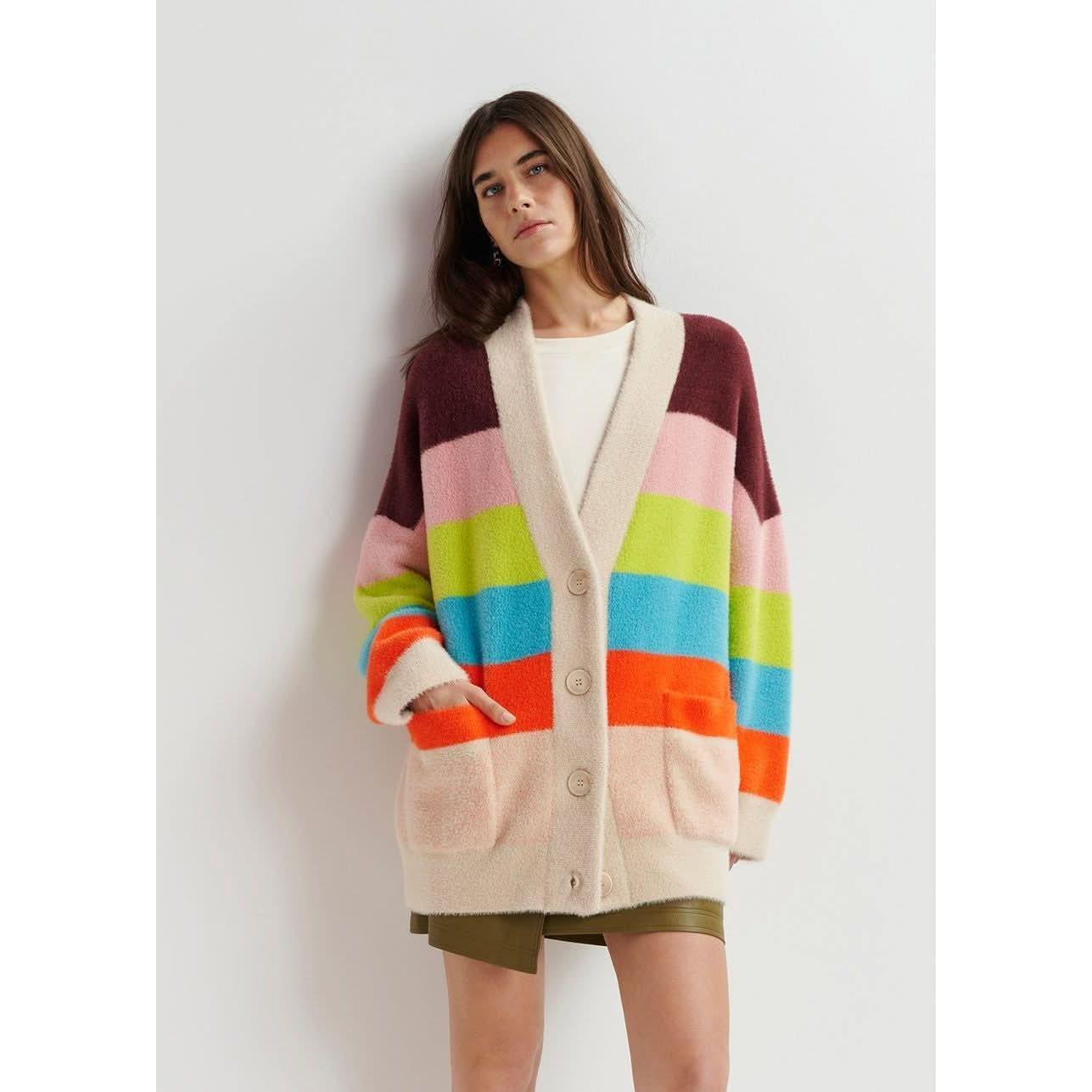 A person stands against a plain white background wearing the Essentiel Antwerp Multicolor Striped Fuzzy Knitted Cardigan, featuring an oversized fit and vibrant horizontal stripes in shades of maroon, pink, green, blue, and orange.