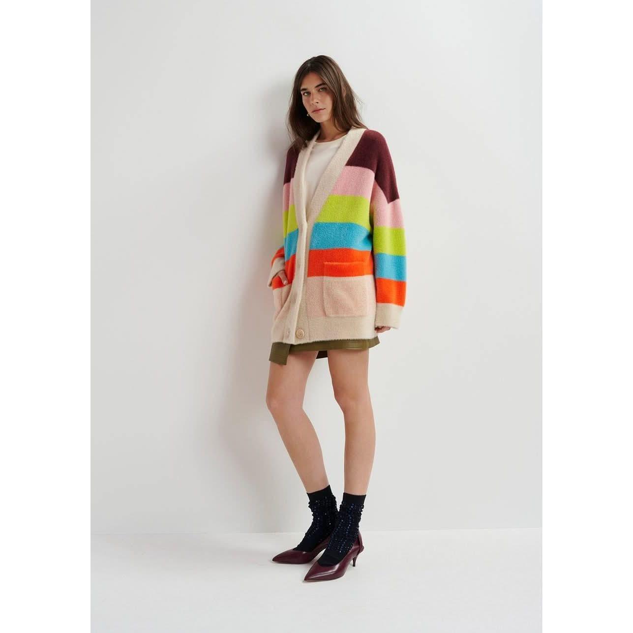 A person wearing the Essentiel Antwerp Multicolor Striped Fuzzy Knitted Cardigan, known for its oversized fit, pairs it with a white shirt and green skirt while standing against a white wall. They complete their casual yet stylish look with dark ankle-length socks and maroon heels.