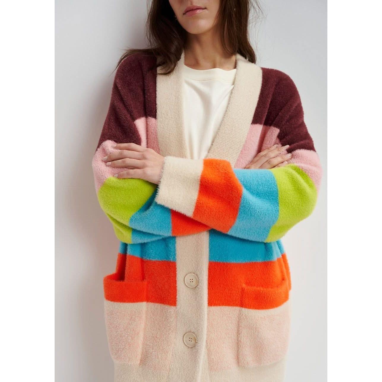 A person wearing the Essentiel Antwerp Multicolor Striped Fuzzy Knitted Cardigan, with an oversized fit and large buttons, showcases shades of pink, brown, green, blue, orange, and beige. The mid-weight knit design from Essentiel Antwerp adds comfort as they stand with crossed arms against a neutral background.