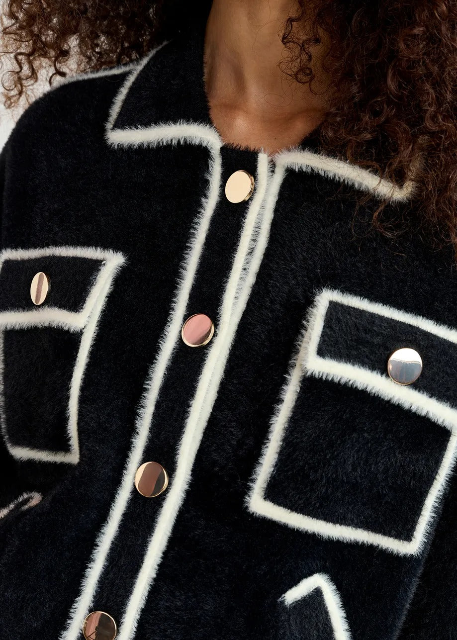 Close-up of the Essentiel Antwerp Galena Cardigan Black, showcasing a black fuzzy fabric with white trim and six gold-tone buttons arranged vertically. The Essentiel Antwerp design, which includes trim outlining geometric shapes, adds a textured and stylish appearance to the mid-weight knit fabric.