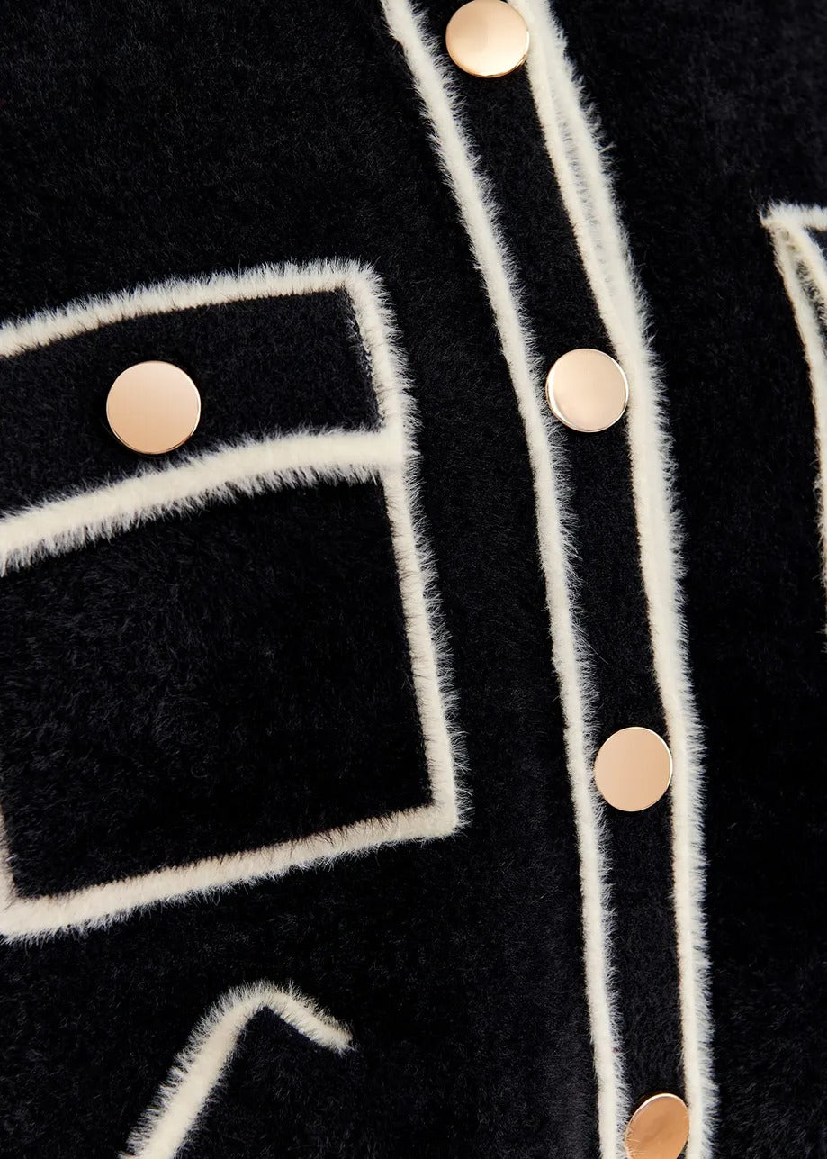 Close-up of the Essentiel Antwerp Galena Cardigan Black, showcasing a black fuzzy fabric with white trim and six gold-tone buttons arranged vertically. The Essentiel Antwerp design, which includes trim outlining geometric shapes, adds a textured and stylish appearance to the mid-weight knit fabric.