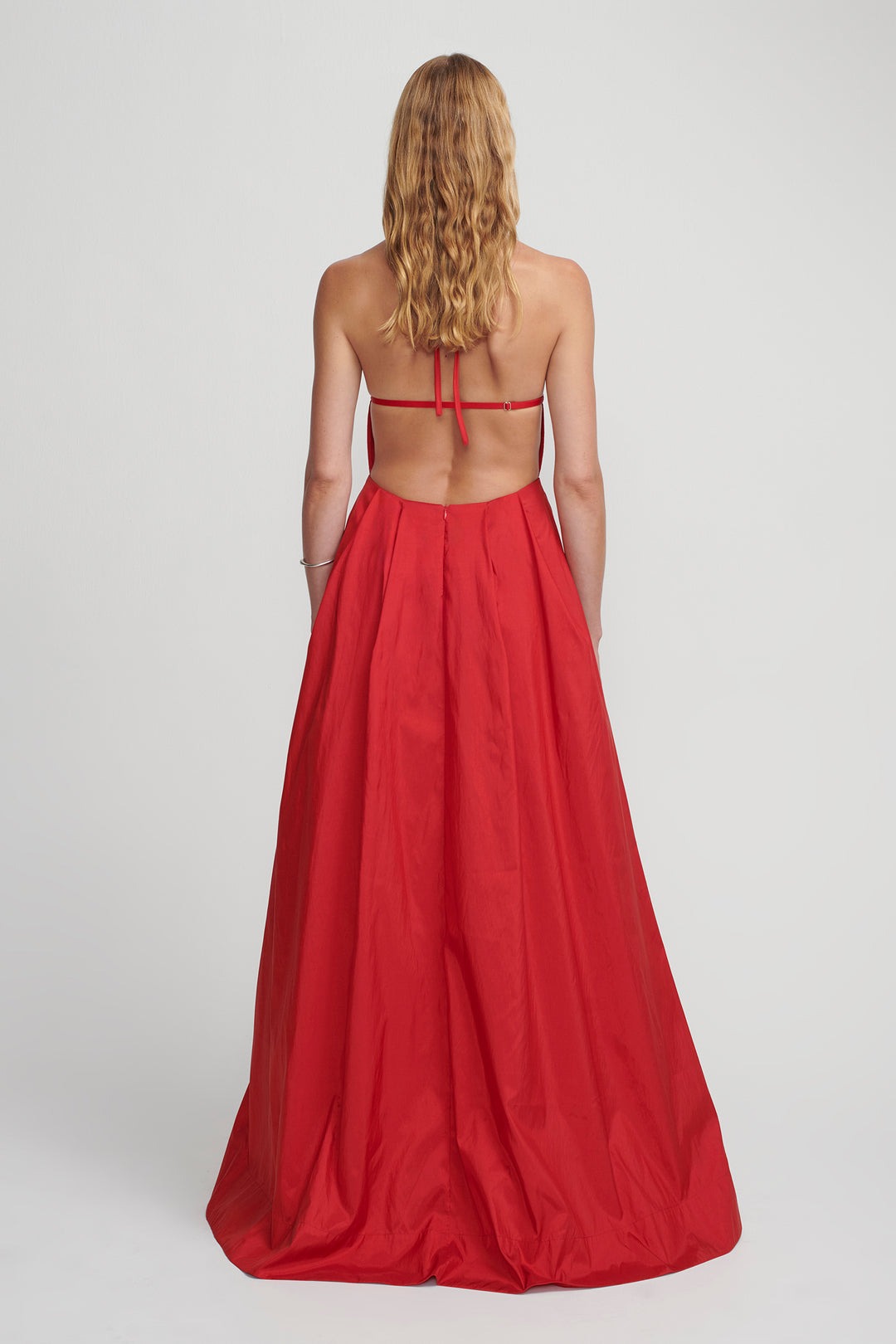 A woman with long, wavy blonde hair is pictured from the back, wearing the Hansen & Gretel Gilda Gown Samba. The strapless red silk maxi dress from Hansen & Gretel features an open back with thin straps tied across her shoulders and a flowing, floor-length skirt. She stands against a plain light gray background.