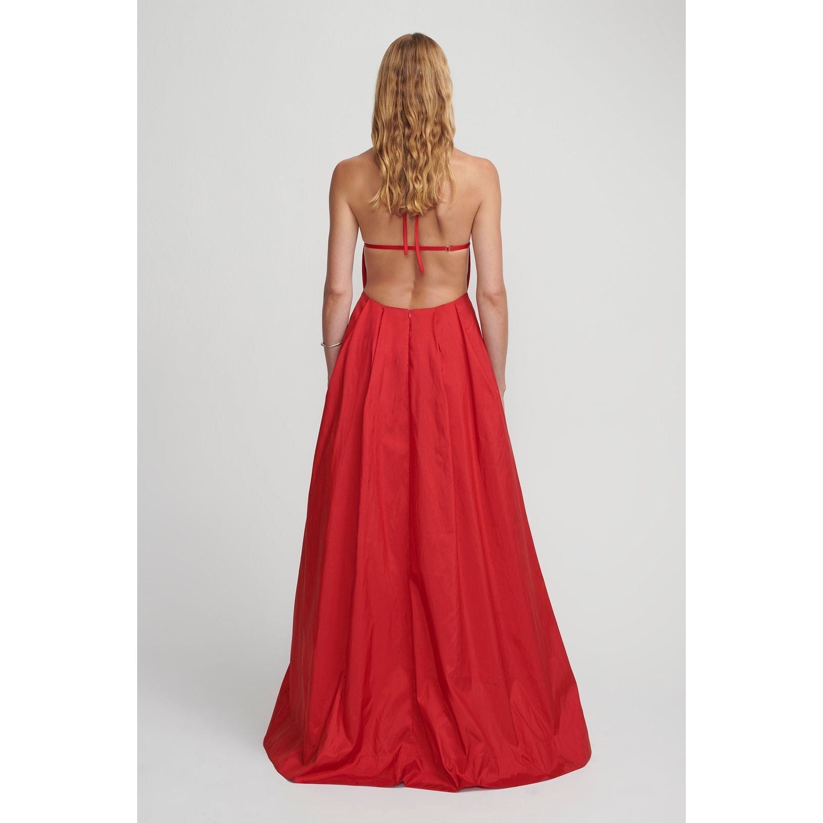 A woman with long, wavy blonde hair is pictured from the back, wearing the Hansen & Gretel Gilda Gown Samba. The strapless red silk maxi dress from Hansen & Gretel features an open back with thin straps tied across her shoulders and a flowing, floor-length skirt. She stands against a plain light gray background.