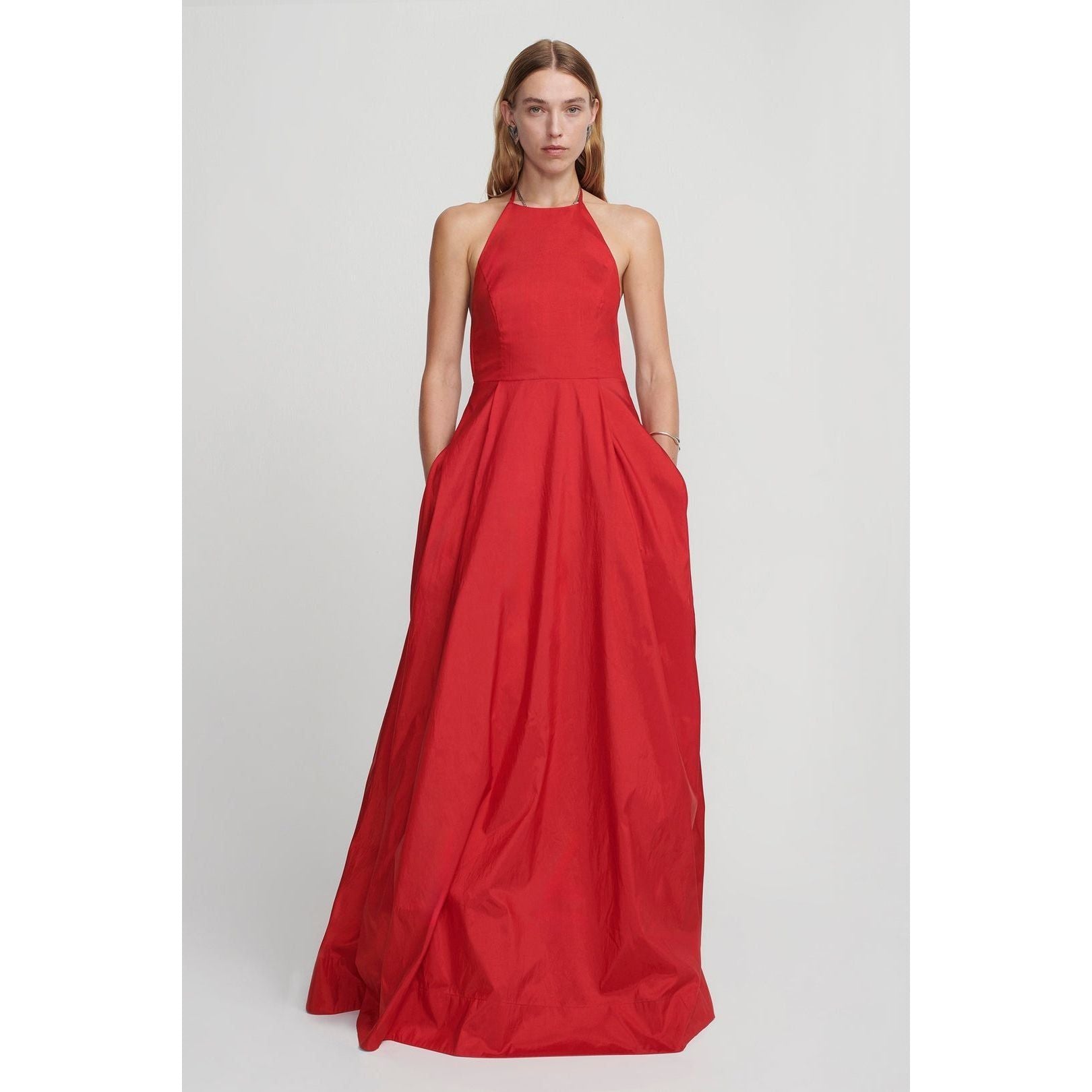 A woman stands against a plain background, wearing the Hansen & Gretel Gilda Gown Samba from Hansen & Gretel. The floor-length red silk maxi dress features a halter neck and a fitted bodice that flares into a full skirt. She has one hand in her pocket and a neutral expression on her face.