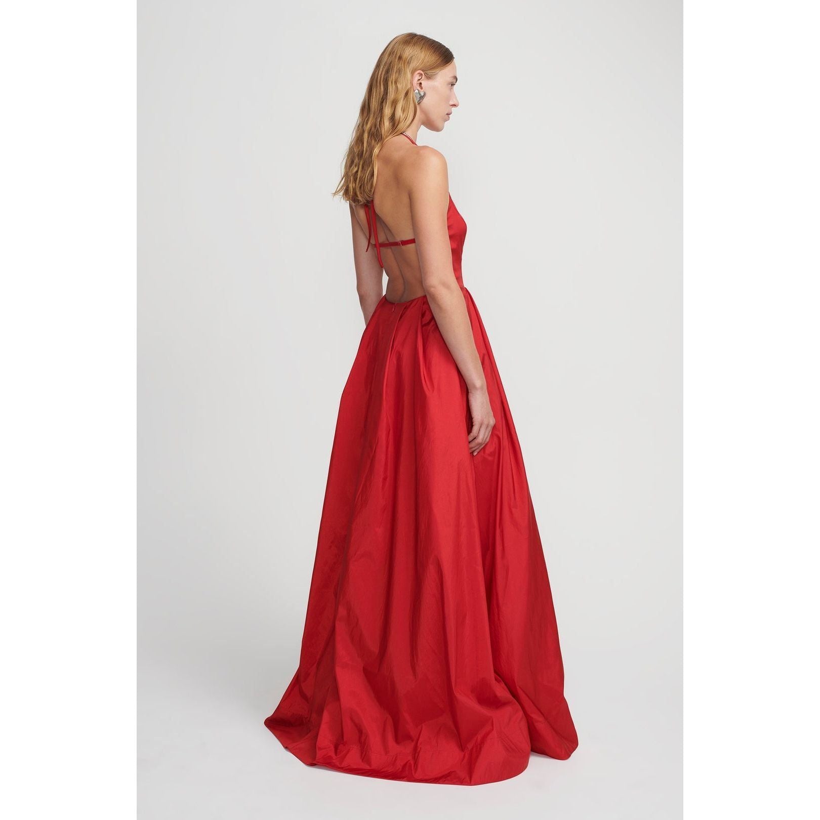 A woman with shoulder-length blonde hair is wearing the Hansen & Gretel Gilda Gown Samba, a floor-length, backless red silk maxi dress with thin straps crossing at the back. She is posed at an angle, revealing the flow and form of the 100% silk gown by Hansen & Gretel. The background is plain, putting full emphasis on the dress.