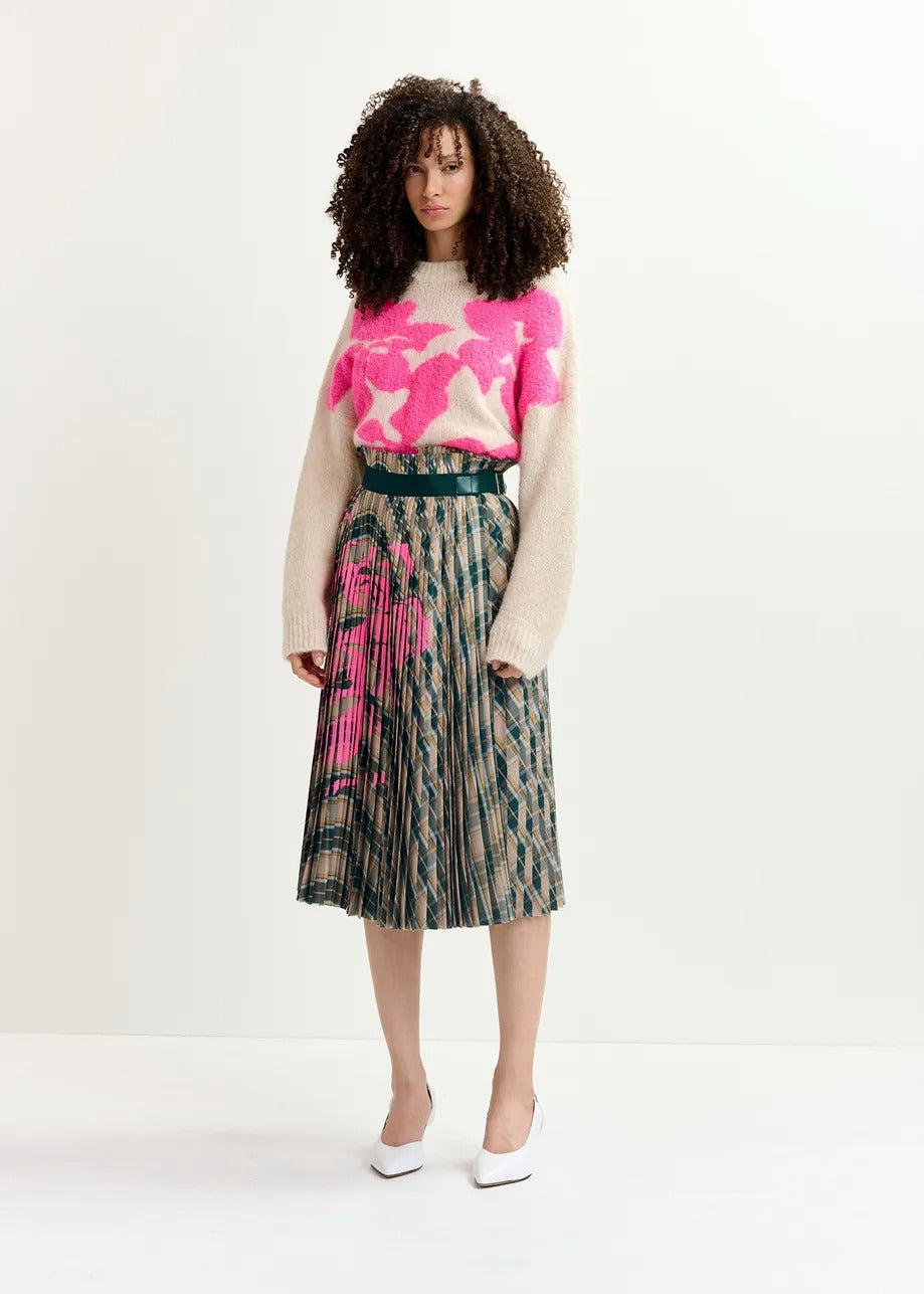 A person with curly hair is wearing a beige sweater with pink and white geometric patterns, paired with the Essentiel Antwerp Godess Skirt—a high-waisted pleated skirt featuring beige and dark green checks along with floral prints. The skirt includes recycled polyester. They are also wearing white shoes, standing against a plain, light-colored background.