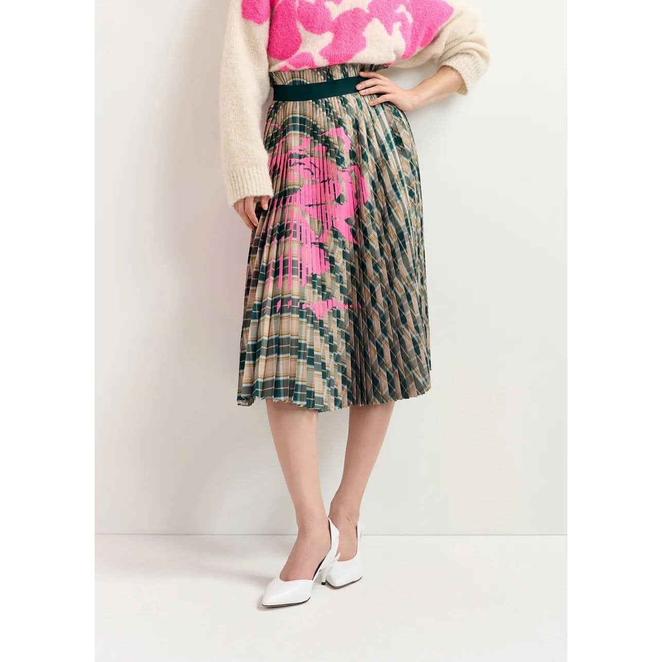 A person is wearing a beige sweater with pink patterns and the Essentiel Antwerp Godess Skirt, a beige and dark green checked pleated skirt with a floral print. They are also sporting white heeled shoes. The image is set against a plain background.