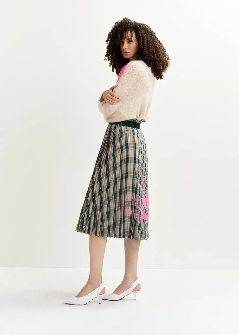 A woman with curly hair stands in profile, wearing a fluffy white sweater and the Essentiel Antwerp Godess Skirt, a beige and dark green checked pleated skirt adorned with a pink floral print. She's also wearing white slingback heels and stands against a plain white background.