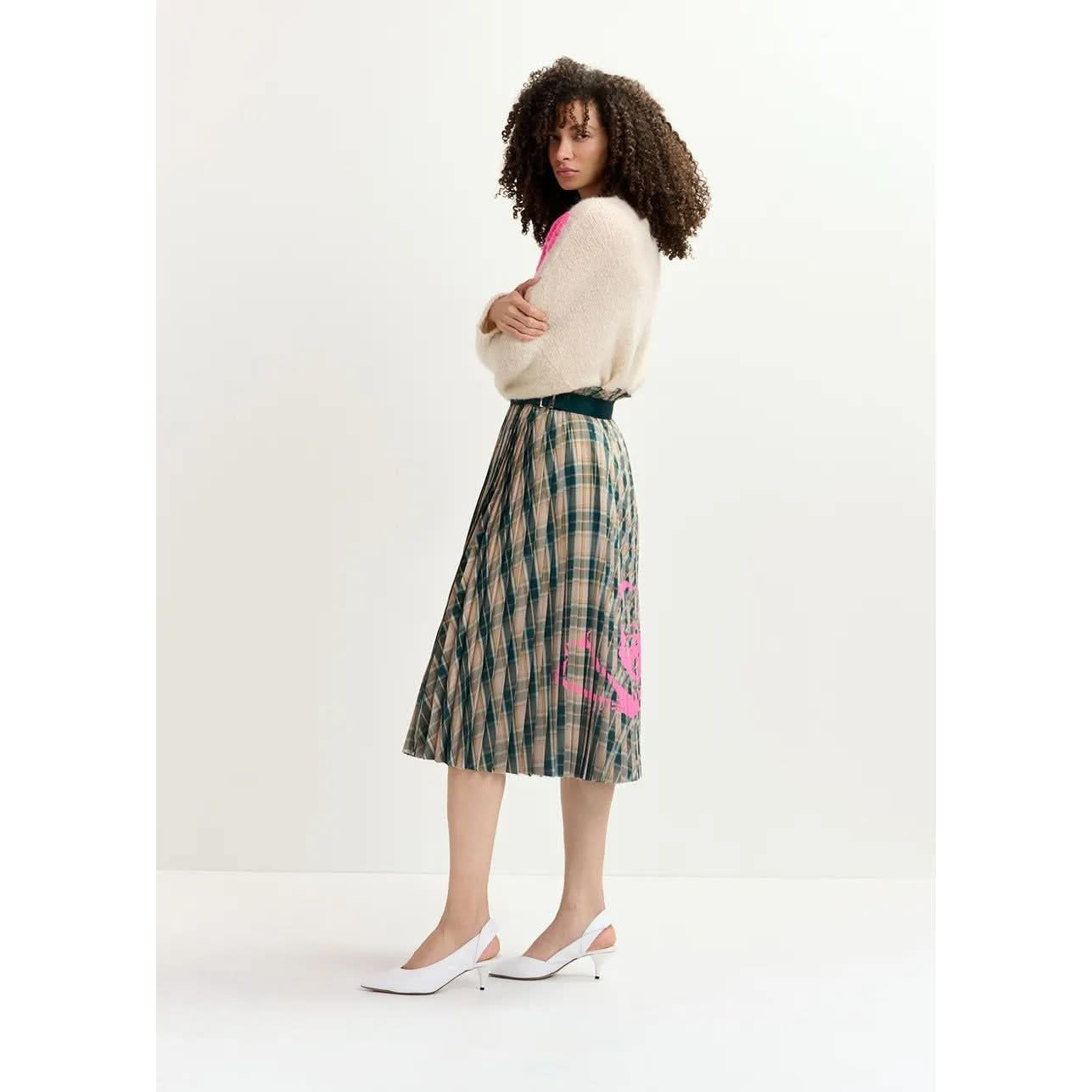 A woman with curly hair stands in profile, wearing a fluffy white sweater and the Essentiel Antwerp Godess Skirt, a beige and dark green checked pleated skirt adorned with a pink floral print. She's also wearing white slingback heels and stands against a plain white background.