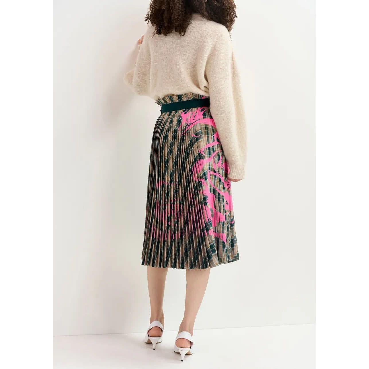 A person with curly hair is facing away, showcasing the back of their outfit. They are wearing a beige, long-sleeved sweater paired with the Essentiel Antwerp Godess Skirt—a high-waisted, pleated skirt featuring a beige and dark green checked pattern adorned with a floral print in shades of green and pink. The ensemble is completed with white heeled shoes.