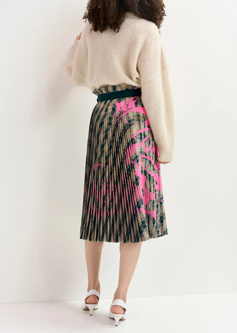 A person with curly hair is facing away, showcasing the back of their outfit. They are wearing a beige, long-sleeved sweater paired with the Essentiel Antwerp Godess Skirt—a high-waisted, pleated skirt featuring a beige and dark green checked pattern adorned with a floral print in shades of green and pink. The ensemble is completed with white heeled shoes.