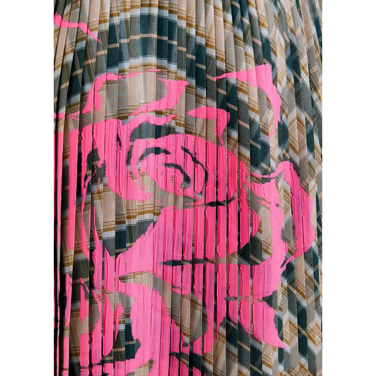 A close-up of the Essentiel Antwerp Godess Skirt reveals its pleated fabric adorned with intersecting plaid lines in various shades of green, brown, and beige. A vivid, abstract pink design overlays the plaid background on this high-rise waistband skirt made from recycled polyester, creating a striking contrast.