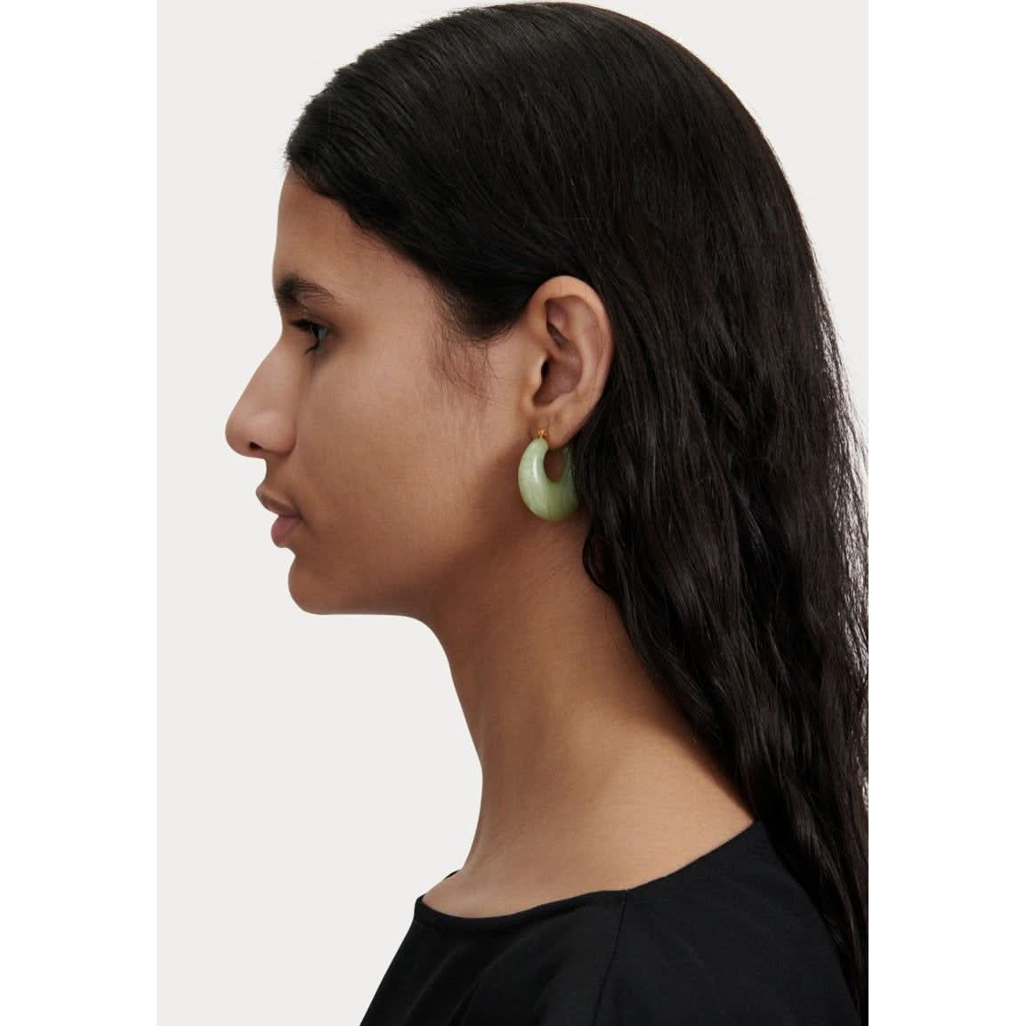 Profile of a person with long dark hair, wearing a black top and striking Rachel Comey Grass Earrings in Pear. The background is plain white.