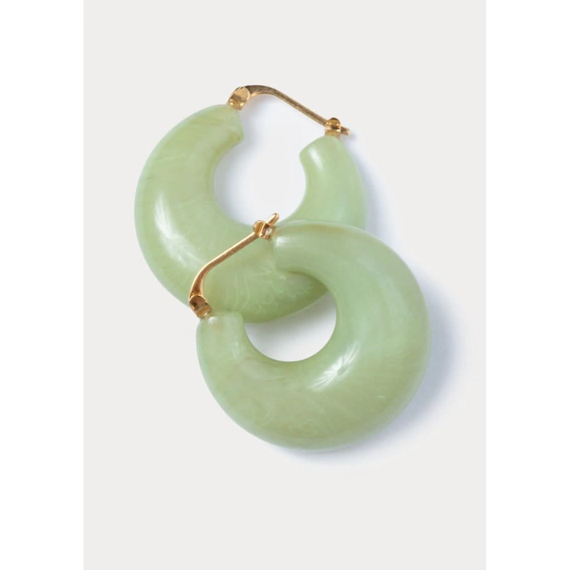 A pair of Rachel Comey Grass Earrings in Pear feature crescent-shaped, translucent green acrylic hoops with gold-toned clasps, elegantly displayed against a plain light background. Crafted in Italy, these earrings boast a smooth finish and an elegant design.