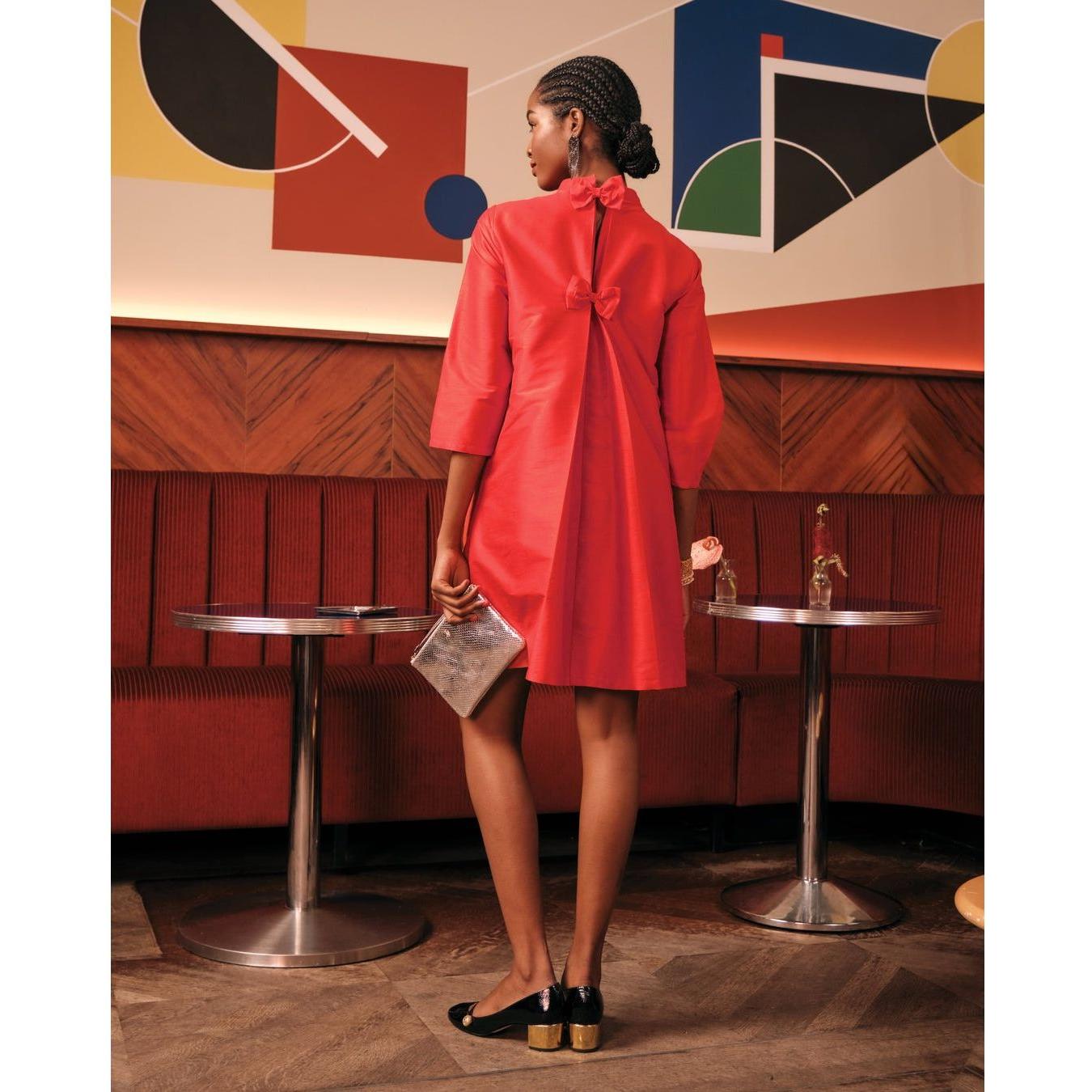 A person wearing the Frances Valentine Diplomat Mini Dress in red stands facing away in a retro-style cafe with geometric art on the wall. They hold a silver clutch and a flower, with two round tables beside them and red upholstered seating.