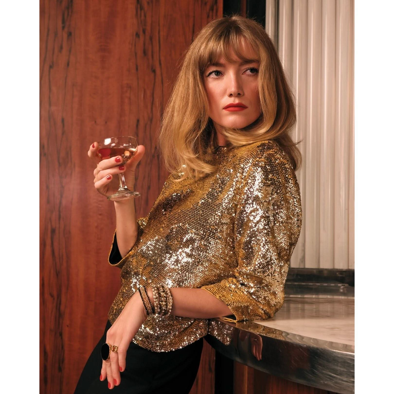 A person with long blonde hair leans against a bar, champagne glass in hand. Wearing a shimmering Frances Valentine Taylor Sequin Top Floral and several bracelets, they sport a contemplative expression. The background, featuring wood and metal accents, complements their chic evening ensemble.