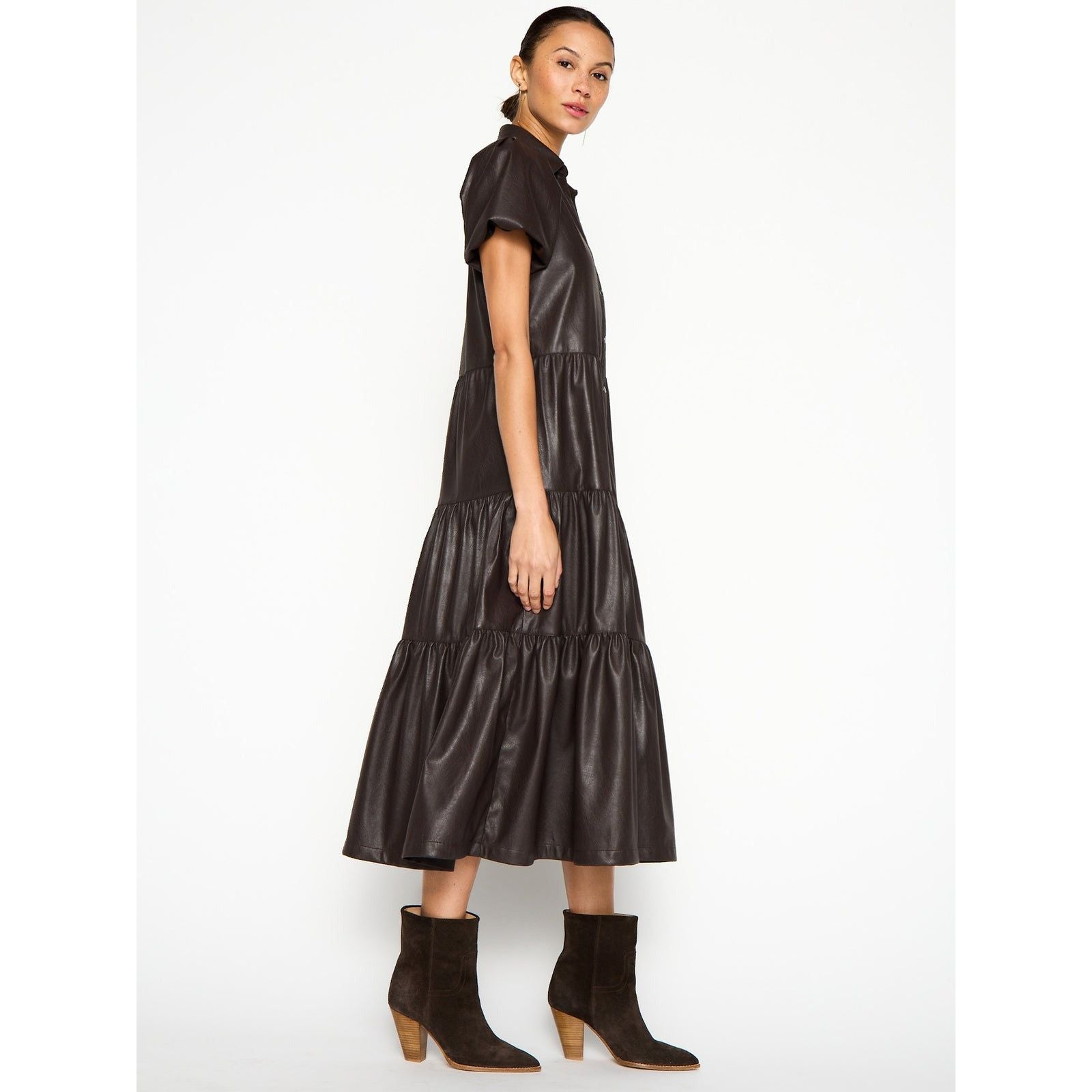 A person is wearing a black, short-sleeved Brochu Walker Havana Dress Vegan Leather with a tiered midi skirt. They are also wearing brown suede ankle boots with chunky heels. The individual has their hair pulled back and is posed in profile against a plain white background.