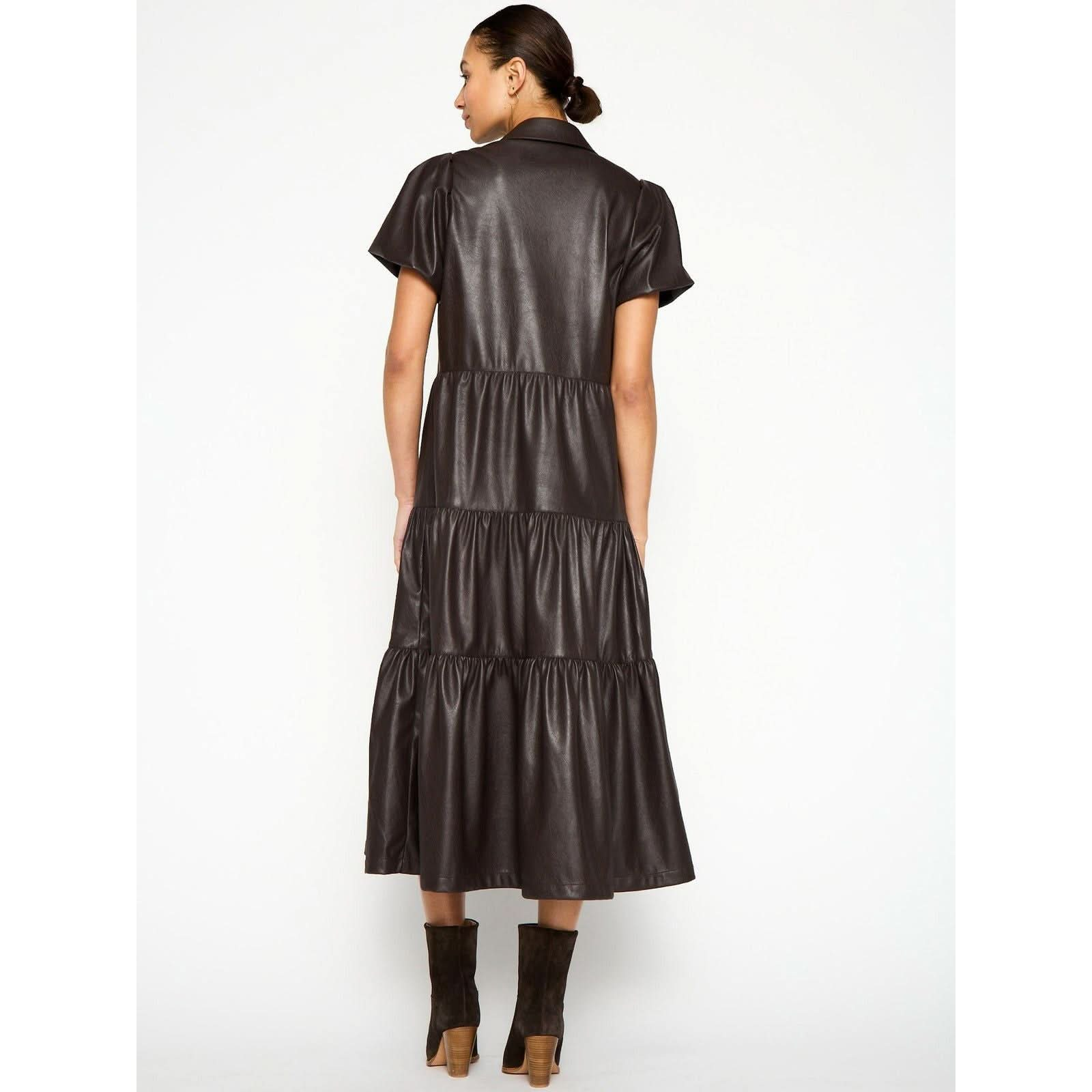 A woman with her hair in a bun is wearing the Brochu Walker Havana Dress Vegan Leather, a short-sleeve, tiered dress that falls mid-calf, paired with brown ankle boots. She is facing away, showcasing the back of this stylish Brochu Walker dress against a plain light background.