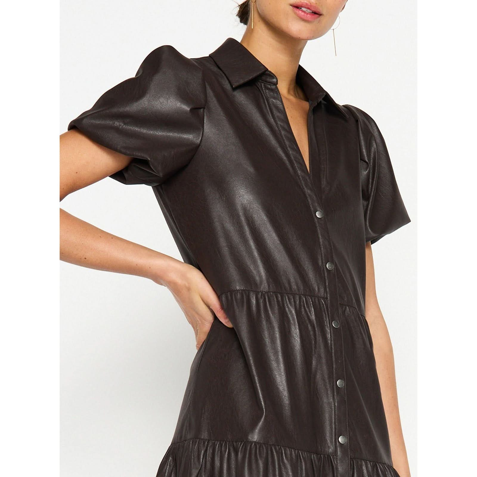 A woman wearing the Brochu Walker Havana Dress in vegan leather, featuring short sleeves, a dark brown hue, metallic buttons, a collar, gathered waist, and puff sleeves. She has one hand on her hip and is partially smiling.