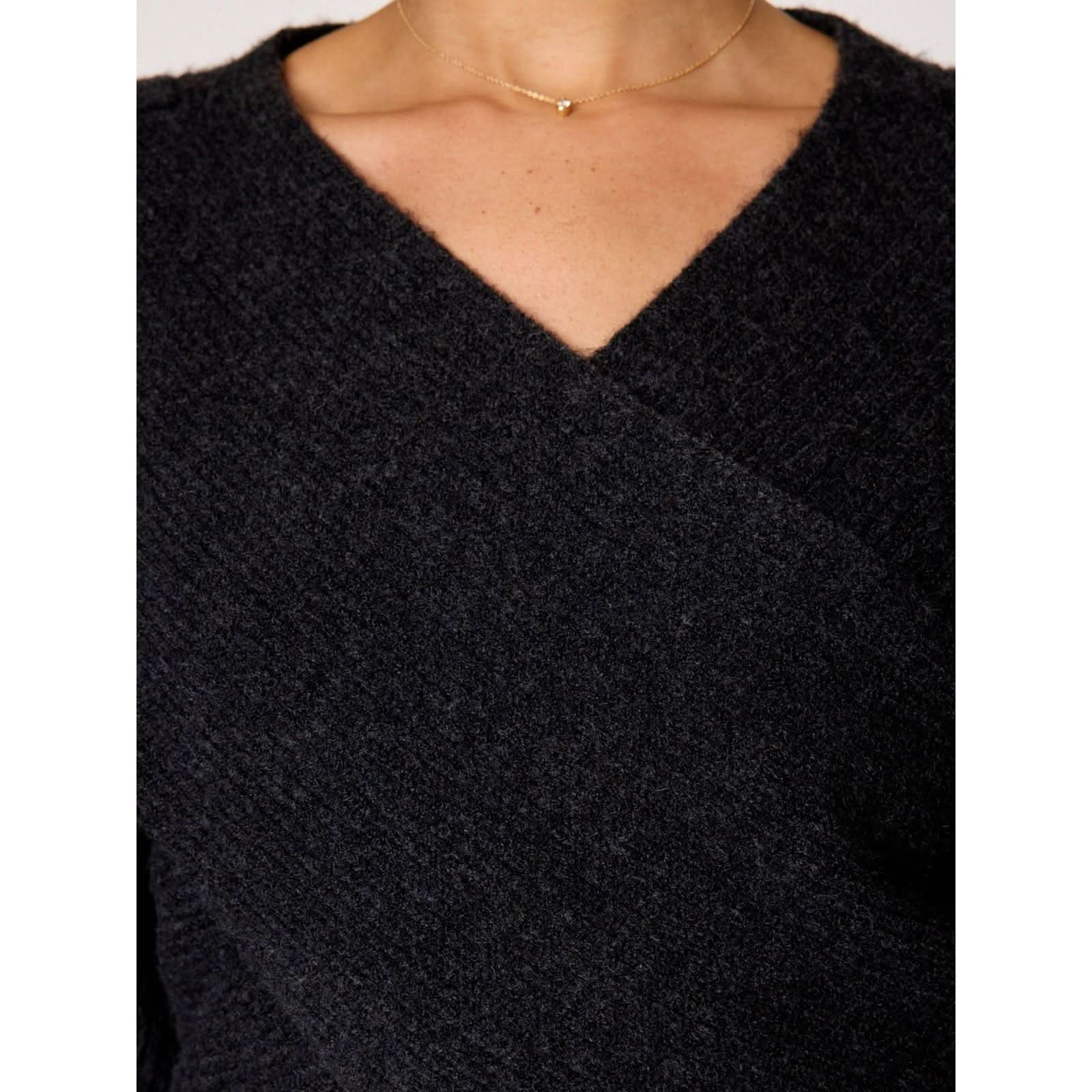 Close-up of a person's chest and neck wearing a black, textured, V-neck top from Brochu Walker's Hughes Wrap Front Sweater Black. The person is also wearing a delicate gold necklace with a small heart-shaped pendant. In the background, an Asher Cardigan made from cotton wool slub yarn can be seen slightly out of focus.