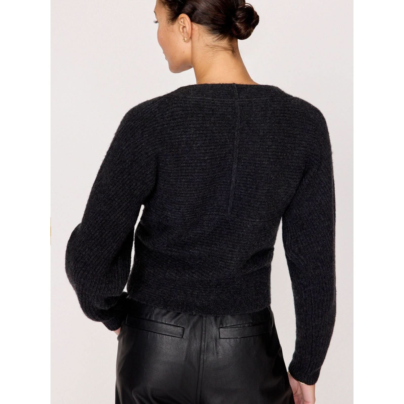 A person with short, dark hair is seen from the back, wearing the Brochu Walker Hughes Wrap Front Sweater in black paired with black leather pants. Their hair is neatly styled in a low bun, and the background is plain and light-colored.