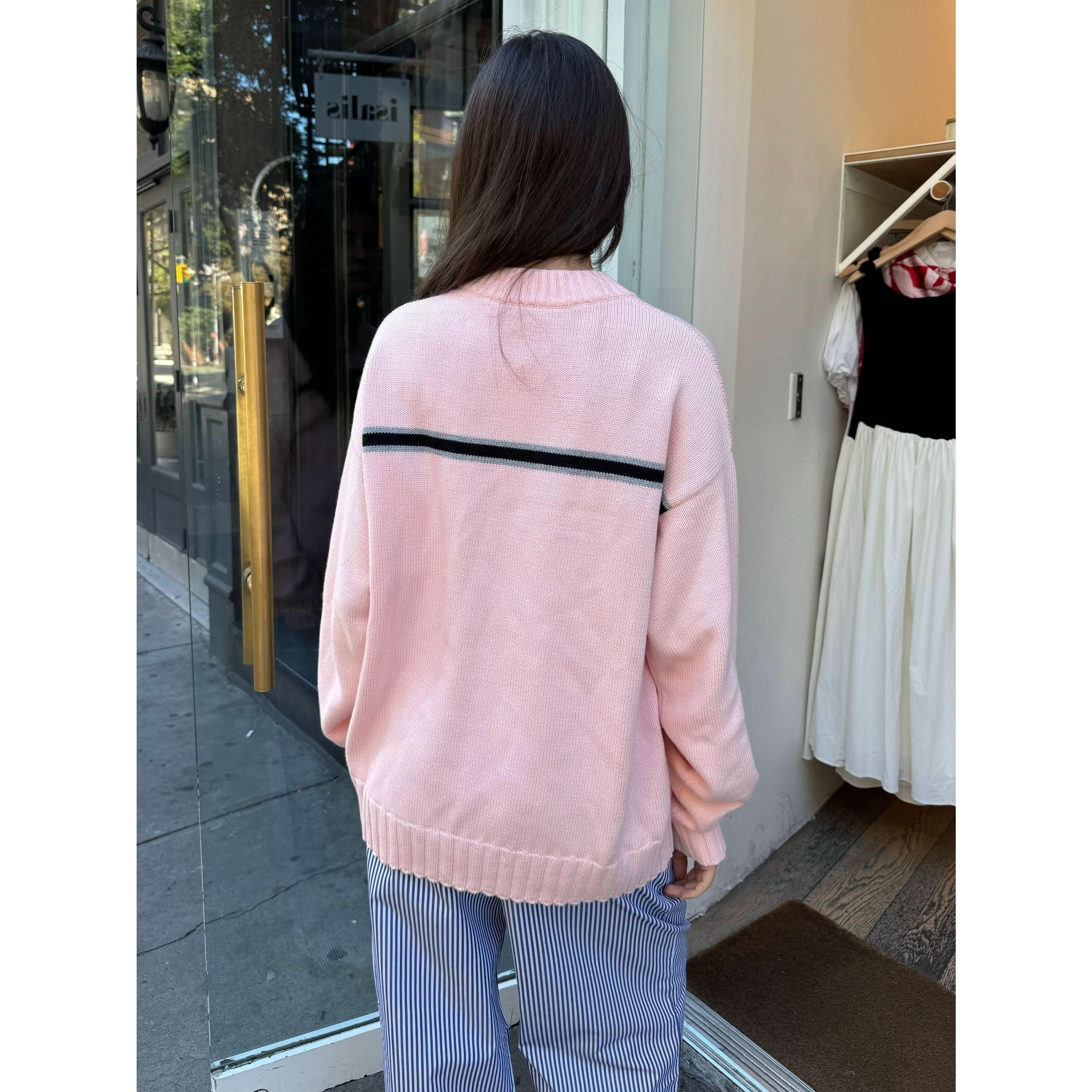 A woman with long brown hair stands outside a store, wearing the Donni The Cotton Crewneck Stripe Sweater featuring a pink hue and a black stripe across the back, paired with blue striped pants. Inside the store, a white dress is visible, enhancing the casual, urban atmosphere.