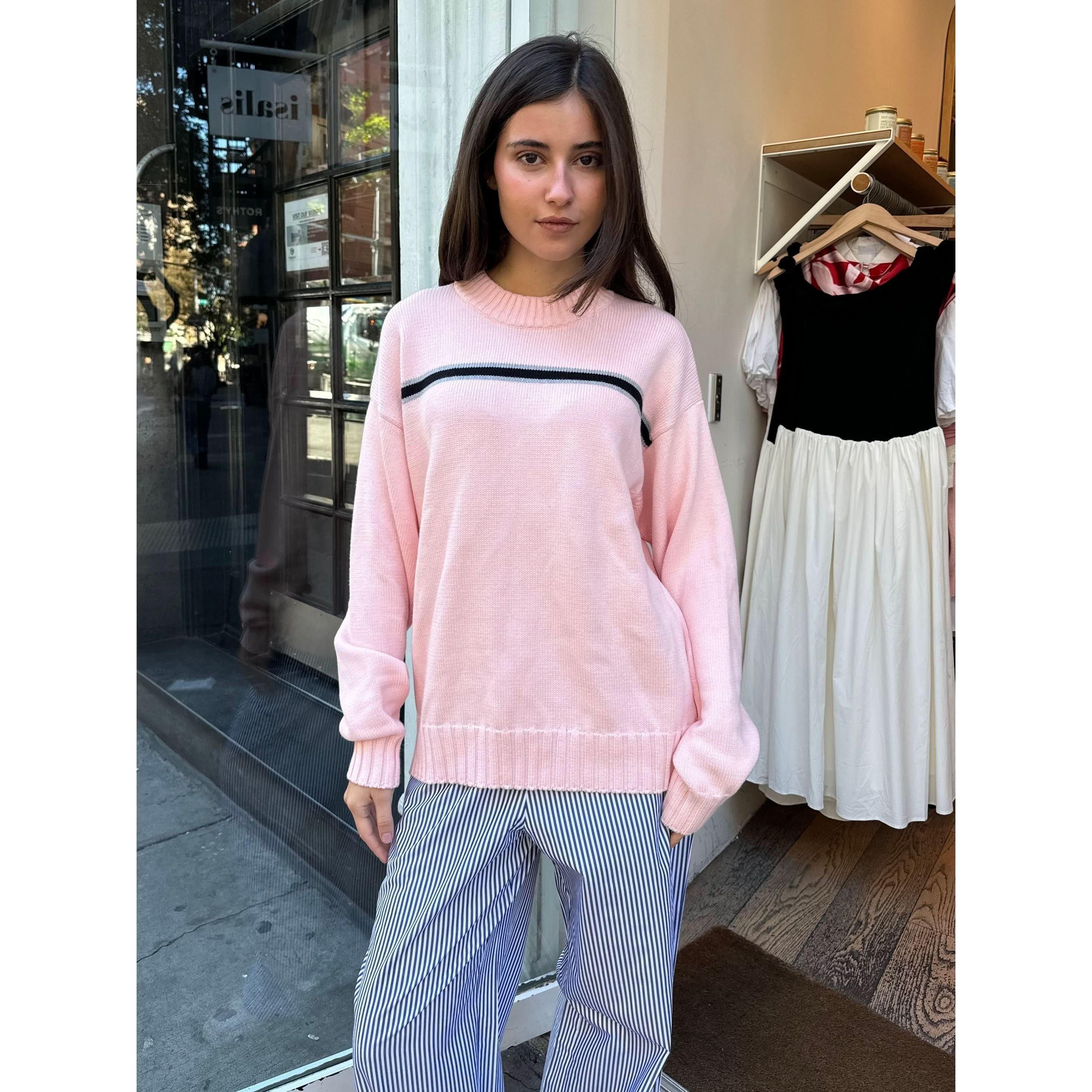 A person with long dark hair stands in a doorway, wearing the Donni The Cotton Crewneck Stripe Sweater in light pink, paired with blue striped pants. A clothing rack with dresses is visible inside the store.