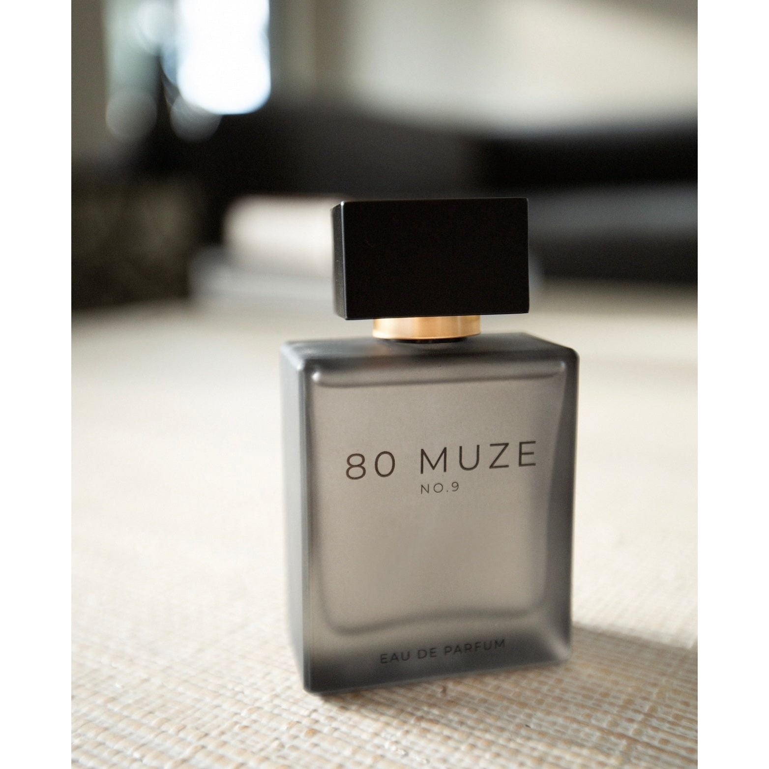 An 80 Muze No. 9 Eau de Parfum bottle rests on a light-colored surface, featuring a sleek rectangular design made from frosted glass and topped with a black cap. The fragrance harmonizes notes of bergamot and green tea, capturing elegance in each spray.