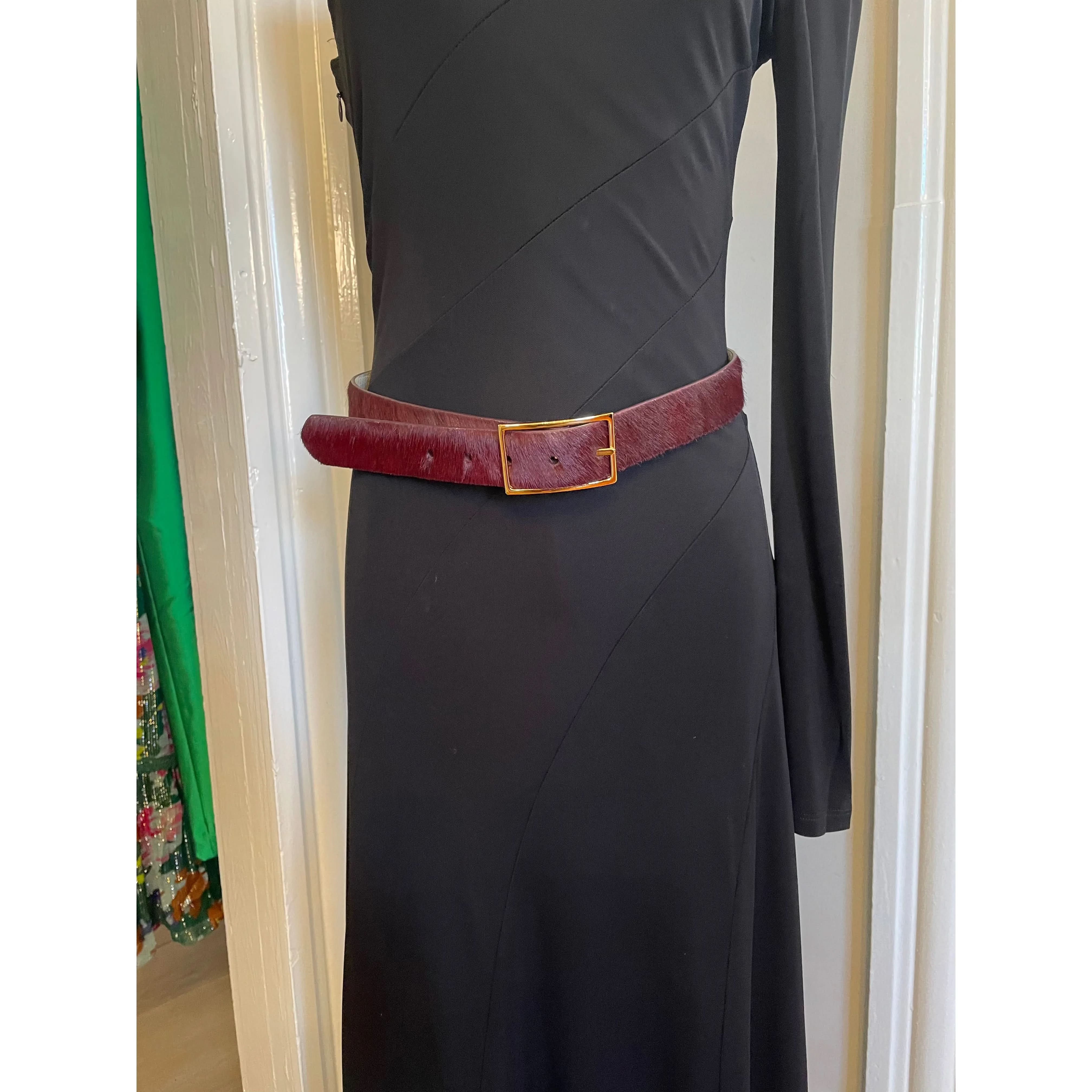 A black dress with long sleeves is accessorized by a W. Kleinberg reversible burgundy and cream calf hair belt, complete with a gold buckle, against the backdrop of a white door.