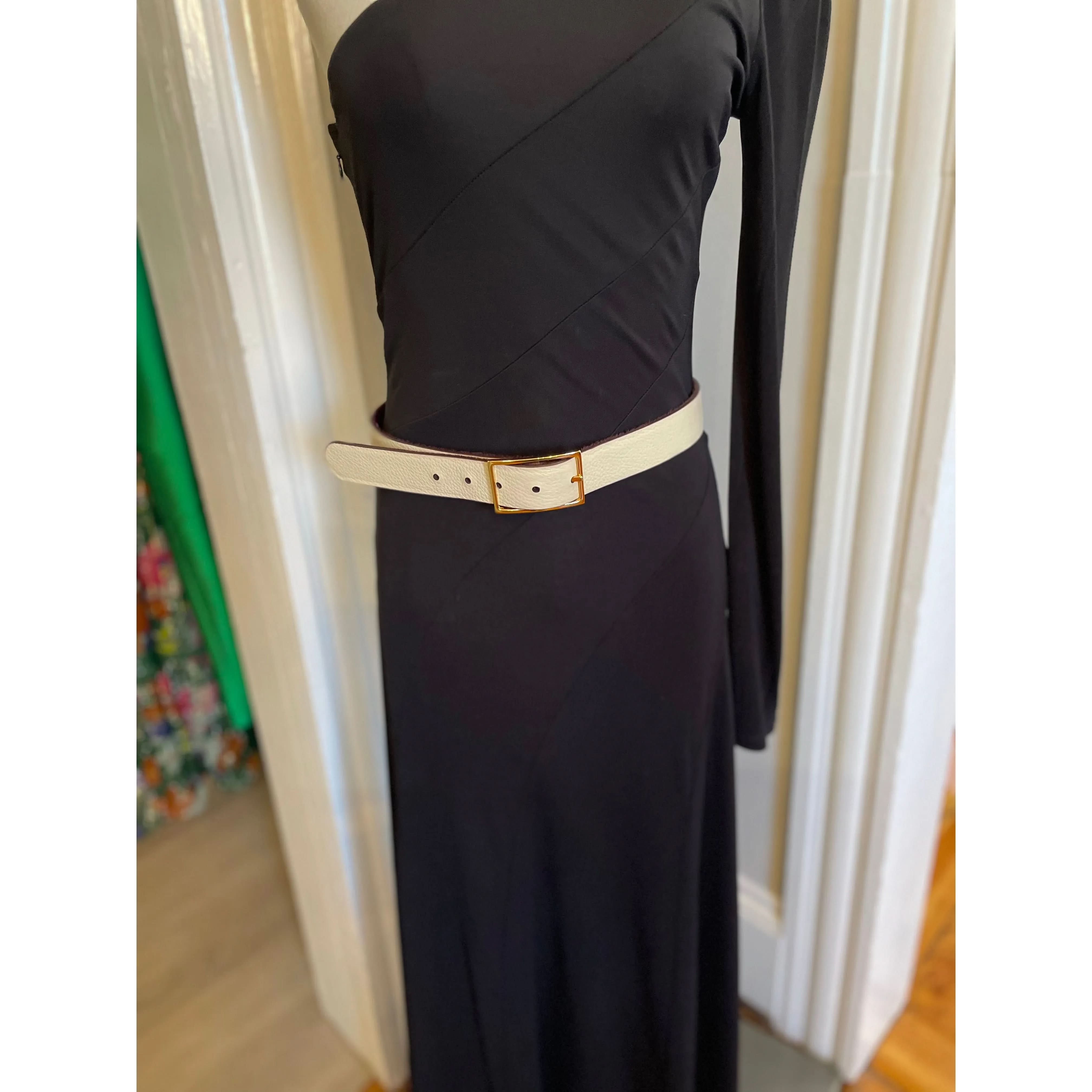 A mannequin displays a black, one-shoulder dress with diagonal seam details, styled with a wide cream pebbled leather belt known as the W. Kleinberg 1 1/4" Hair Calf Reversible Burgundy and Cream with Gold Buckle. The background features white and green door frames.