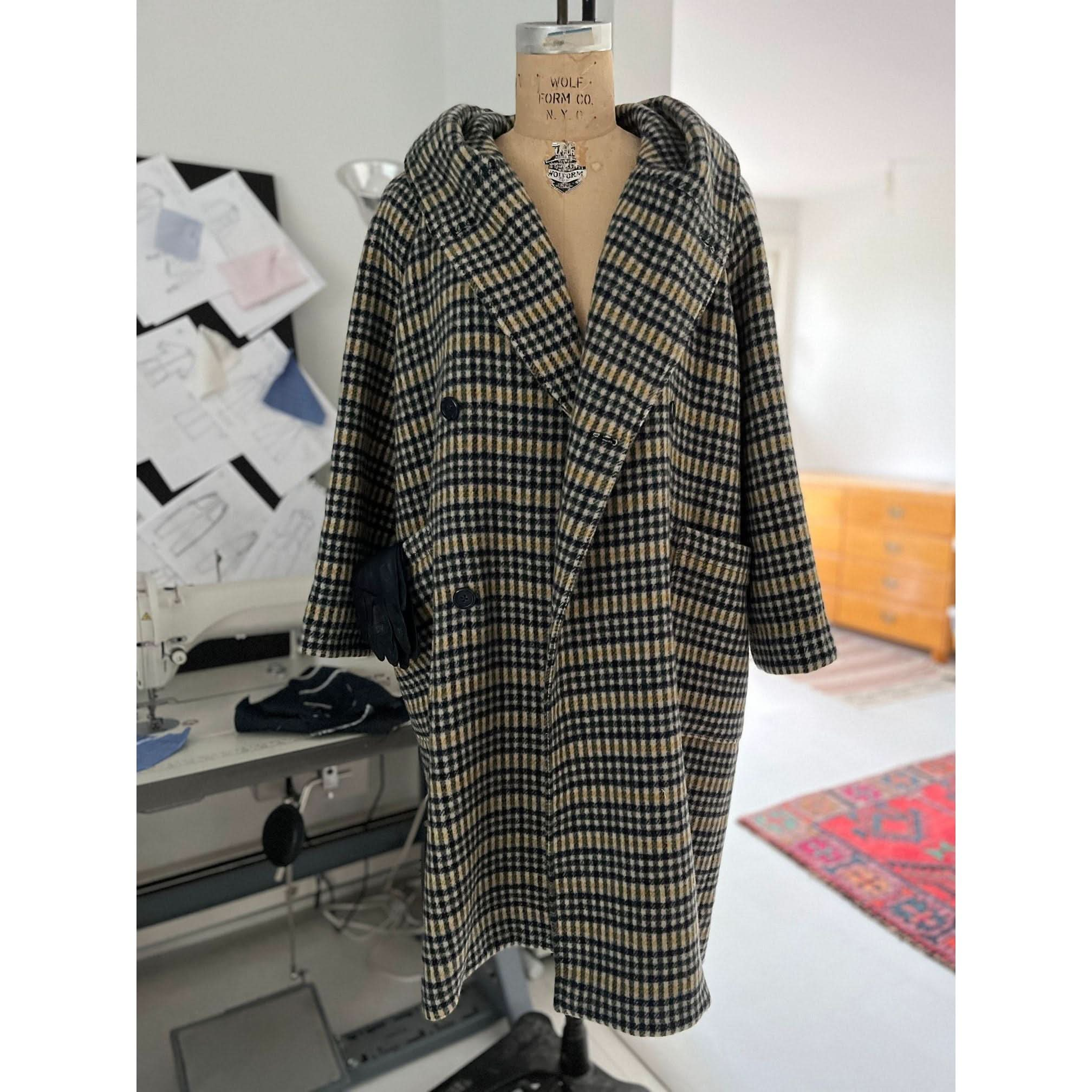 The Heather Harlan Hooded Blanket Coat, skillfully crafted from recycled Italian wool, is showcased on a mannequin in a design studio. The backdrop features sketches, a sewing machine, and a wooden dresser, complemented by a colorful rug on the floor that embodies creative chaos.