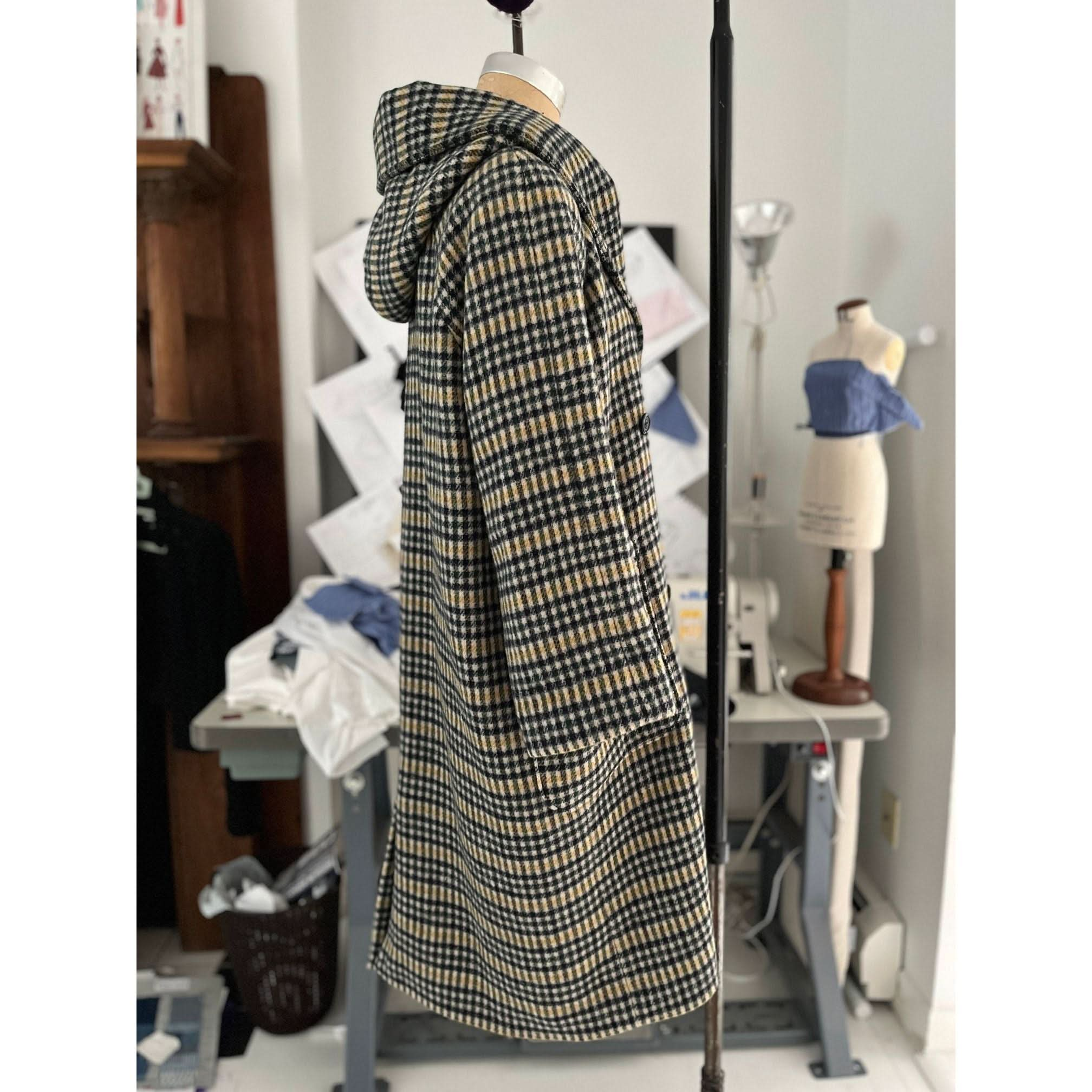 The Heather Harlan Hooded Blanket Coat by Heather Harlan, made from recycled Italian wool, is showcased on a mannequin in a sewing studio. Surrounding it are sewing equipment, fabric, and a dress form adorned partly in blue material, drawing attention to the oversized draped hood that lends an elegant touch.