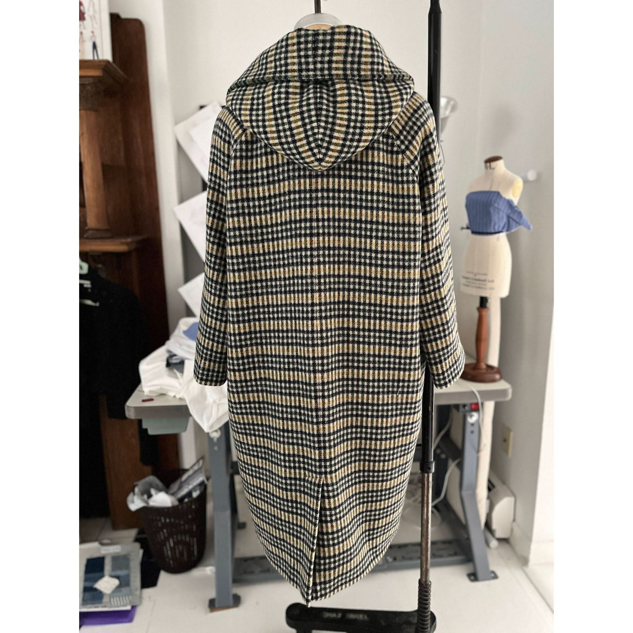 The Heather Harlan Hooded Blanket Coat, crafted from recycled Italian wool, is showcased on a stand featuring its oversized draped hood. In the background, a sewing mannequin, stacks of fabric, sewing patterns, and various sewing room items can be seen. Its plaid design beautifully combines black, beige, and white colors.