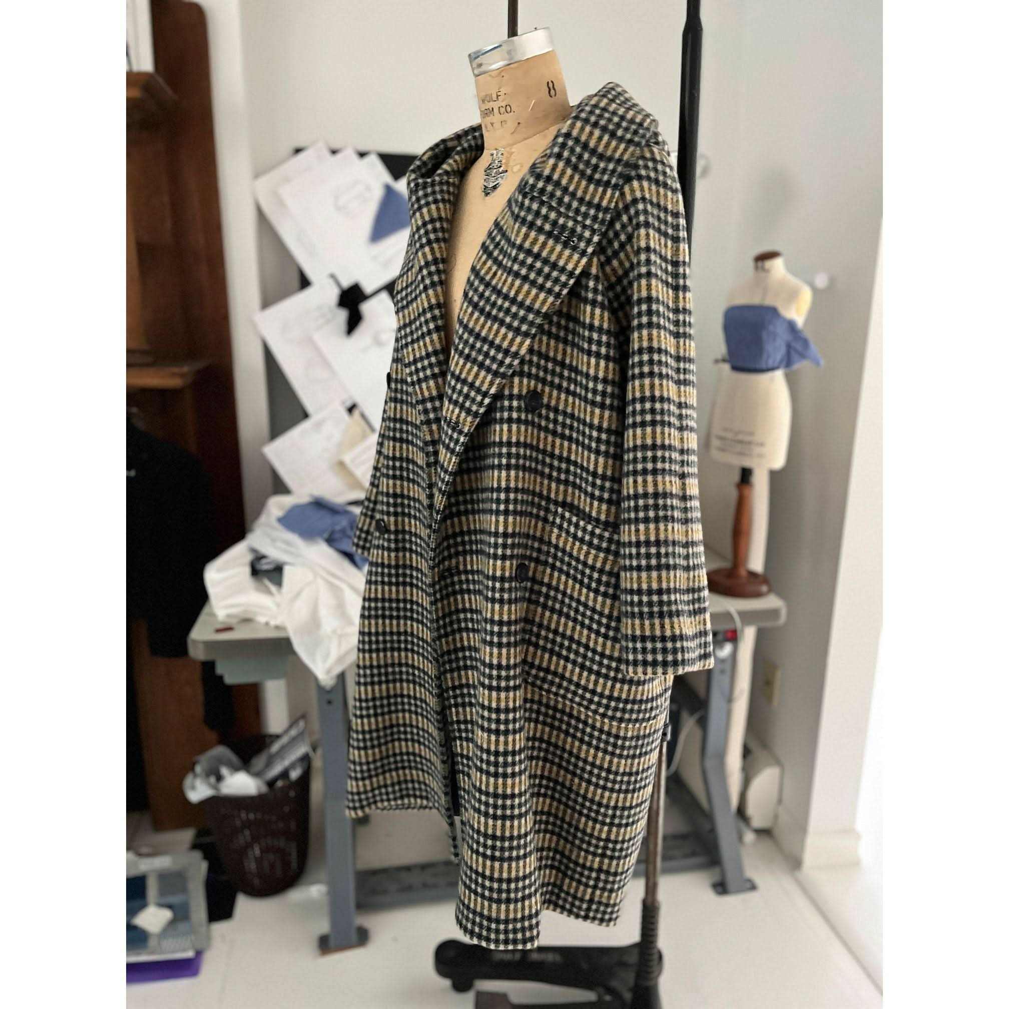 The Heather Harlan Hooded Blanket Coat, created by Heather Harlan and made from recycled Italian wool, showcases a plaid pattern in navy and beige. Displayed on a dress form in a sewing room, it features a wide lapel with an oversized draped hood. The scene also includes a mannequin, pattern papers, and sewing materials.